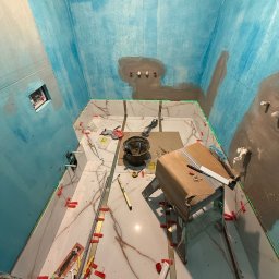 GRCZDR - Łazienka w trakcie układania płytek podłogowych z efektem marmuru, ściany przygotowane z hydroizolacją w odcieniach błękitu, widoczne narzędzia i poziomica laserowa.