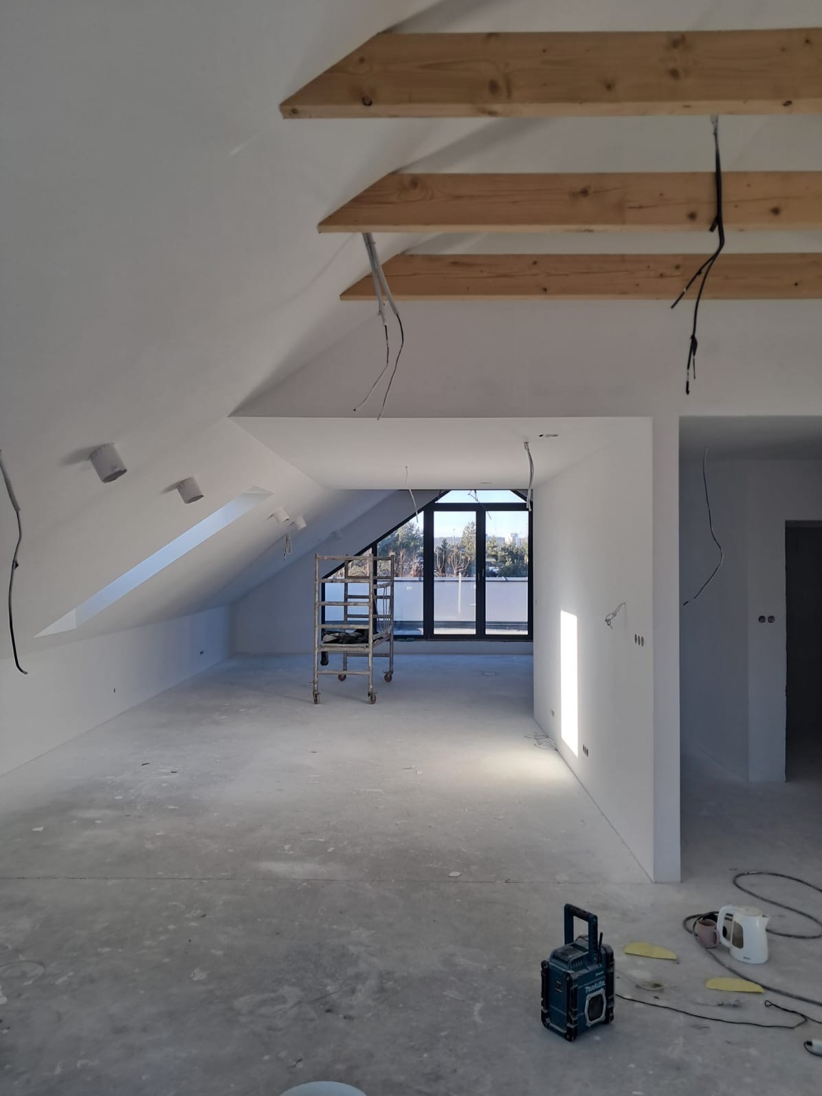 Pokój na poddaszu w trakcie prac wykończeniowych, widoczne drewniane belki stropowe, ściany przygotowane pod malowanie, aluminiowy ruszt oraz lampa budowlana.