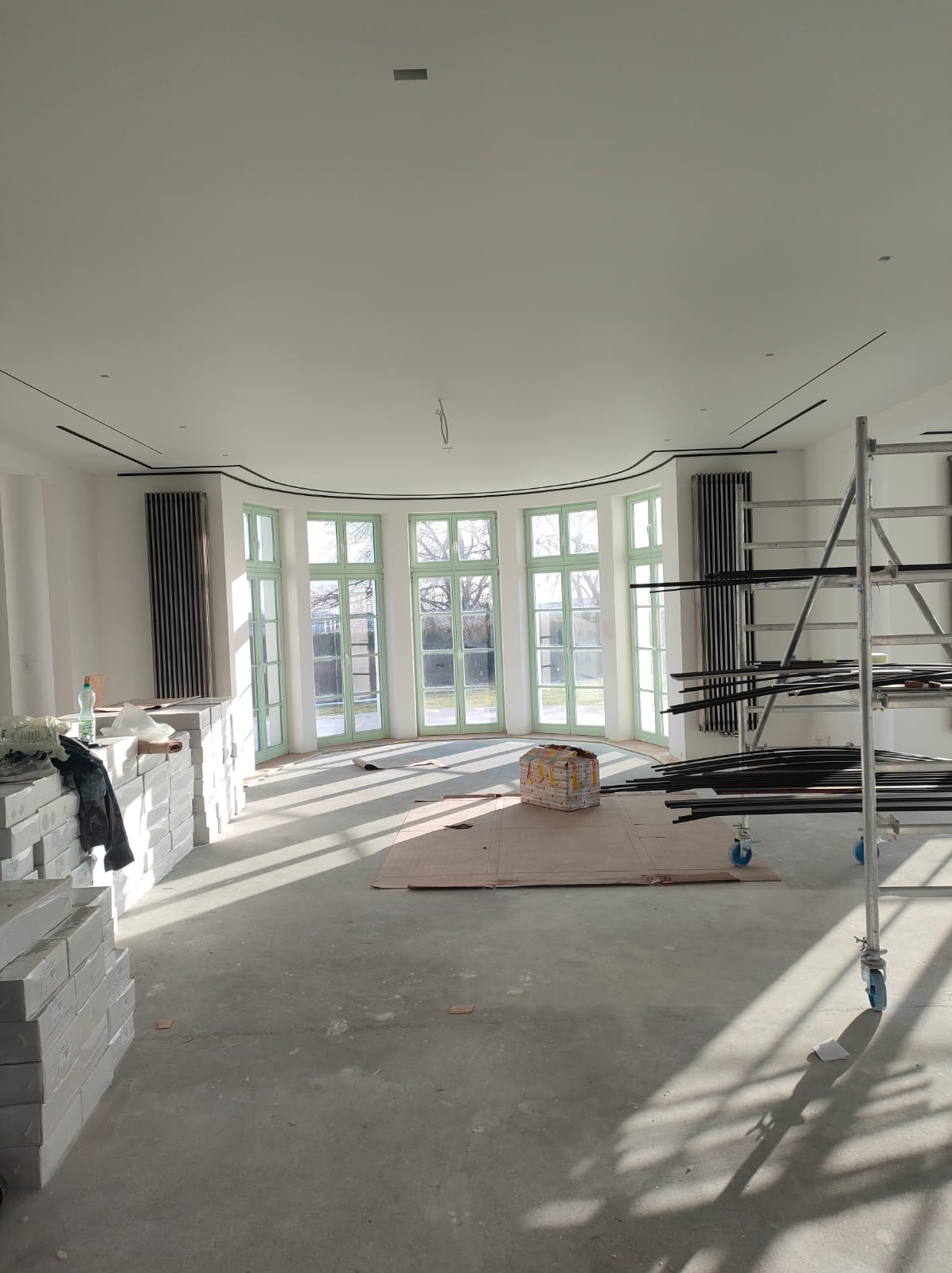 Przestronne wnętrze w trakcie remontu: widoczne cegły, rusztowanie, kartony na podłodze i duże okna z zielonymi ramami wpadające do środka promieniami słońca.
