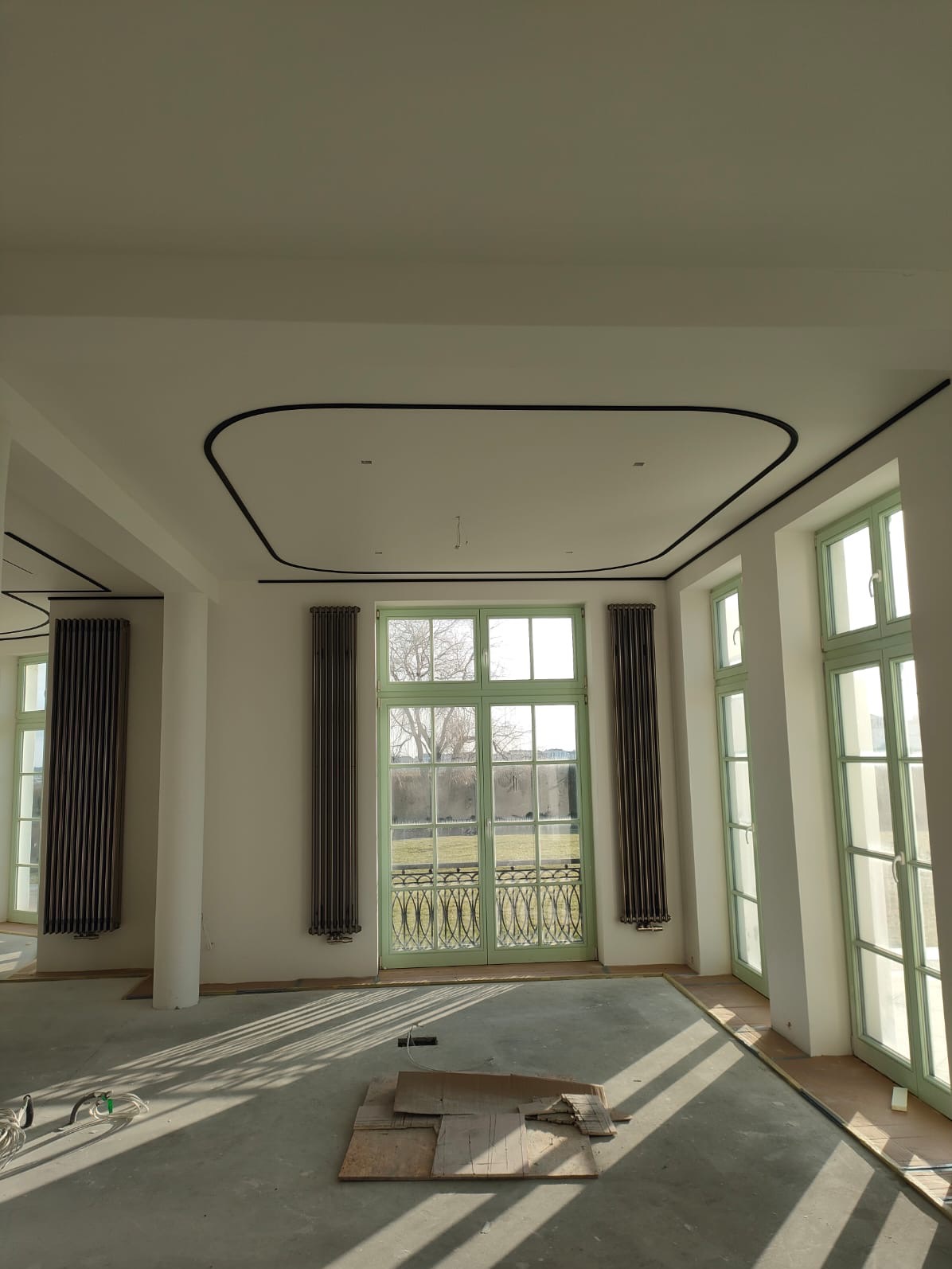 Przestronne wnętrze z wysokimi oknami w zielonych ramach, dekoracyjnym profilem sufitowym i grzejnikami żeberkowymi, surowa podłoga w trakcie prac wykończeniowych.
