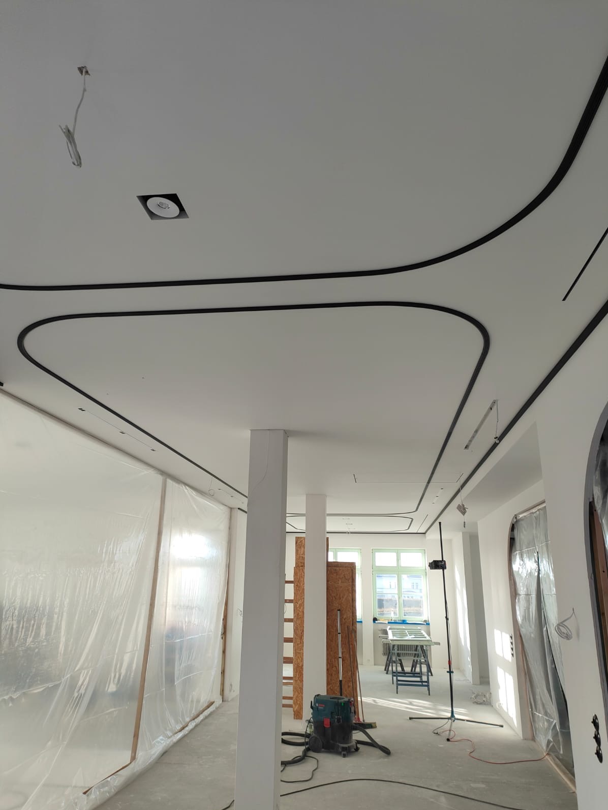 Wnętrze w trakcie prac wykończeniowych: biały sufit z czarnymi, dekoracyjnymi listwami, zakryte folią okna i ściany, widoczny sprzęt budowlany, przygotowanie do montażu oświetlenia.