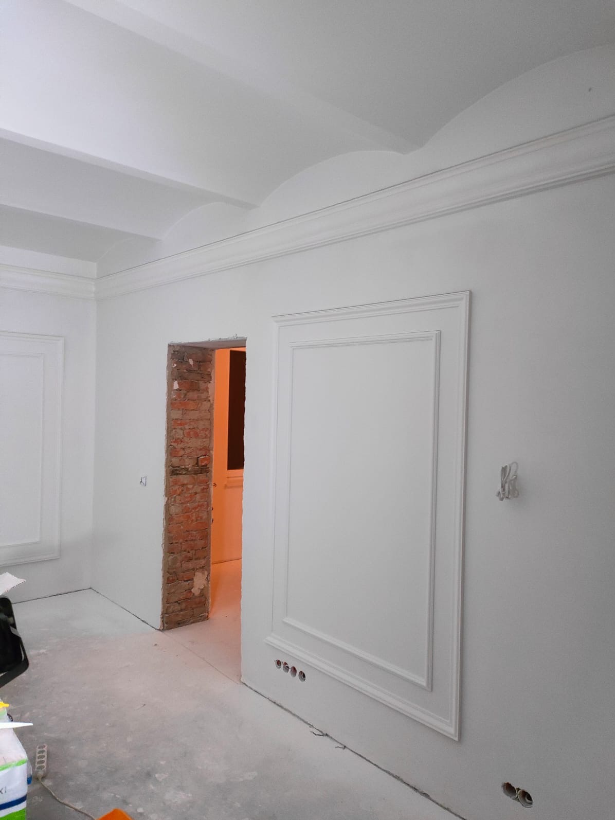 Pokój w trakcie wykańczania z dekoracyjnymi sztukateriami na ścianach i suficie, widoczne fragmenty cegieł wokół otworu drzwiowego, szpachlowana podłoga.