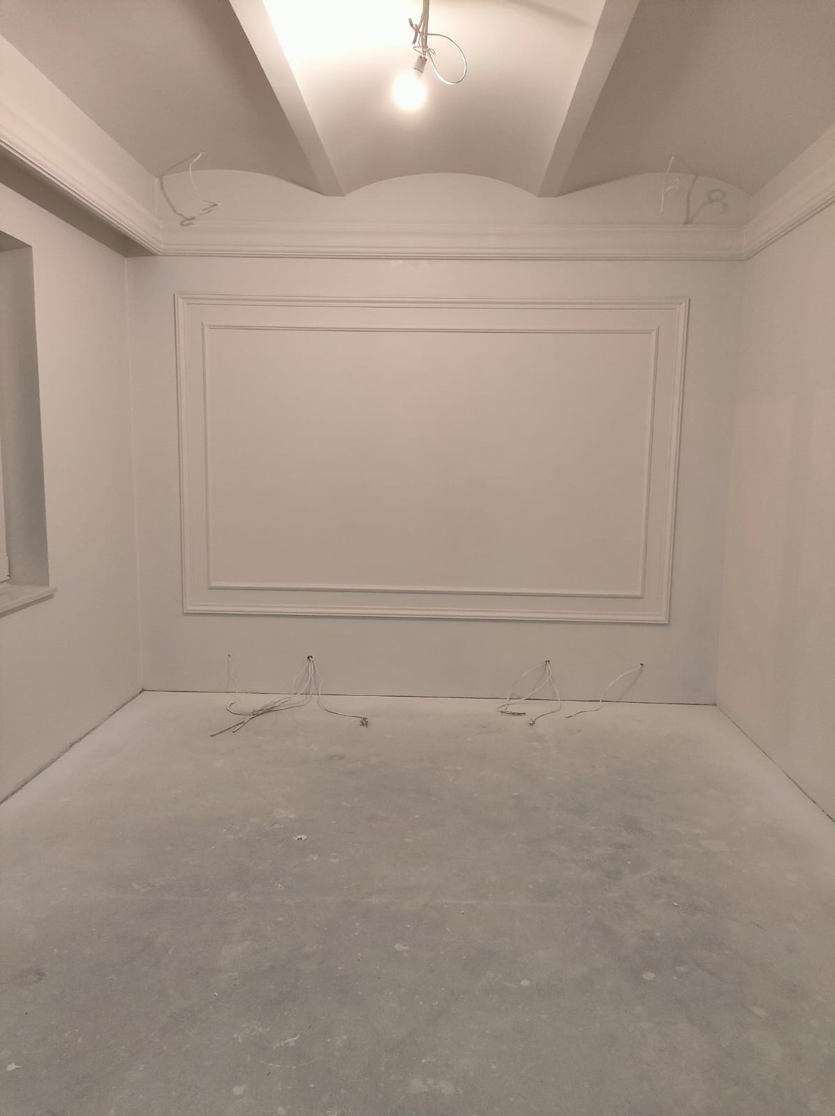 Pusty, biały pokój w trakcie remontu. Na ścianach widoczne dekoracyjne listwy i gzymsy, pod sufitem wystające kable elektryczne. Podłoga pokryta warstwą szpachli lub gładzi. Centralnie umieszczona...