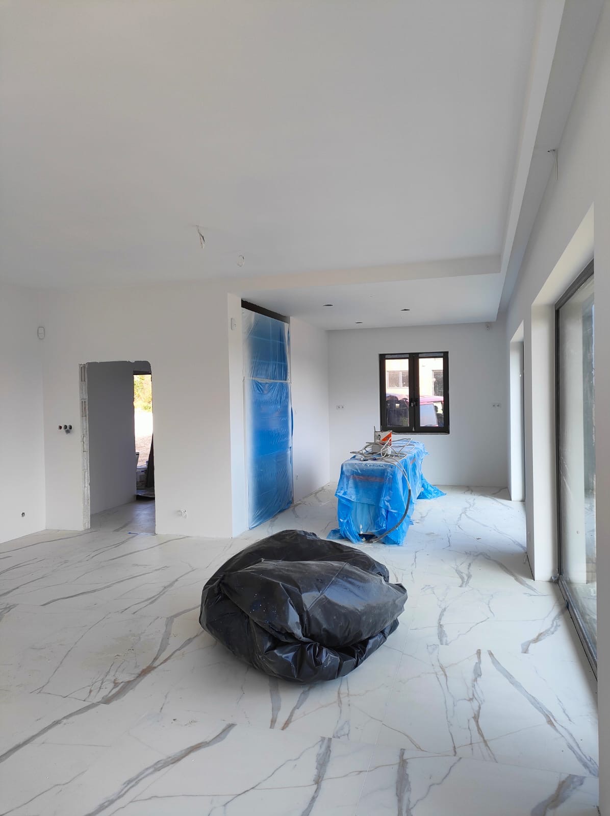 Wnętrze w trakcie prac wykończeniowych: białe ściany, marmurowe płytki podłogowe, zabezpieczone folią drzwi i stół z narzędziami, worek z odpadami na pierwszym planie.