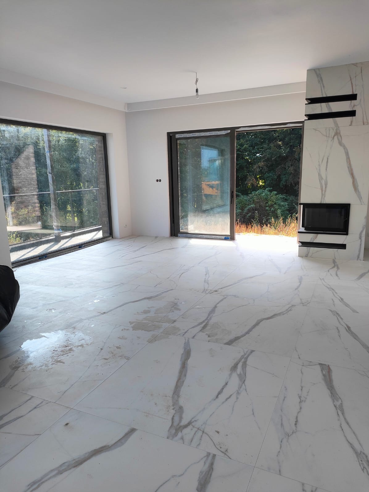 Jasne płytki podłogowe imitujące marmur w przestronnym, nowoczesnym salonie z dużymi oknami i kominkiem obłożonym tym samym materiałem. Widoczne ślady po pracach wykończeniowych.