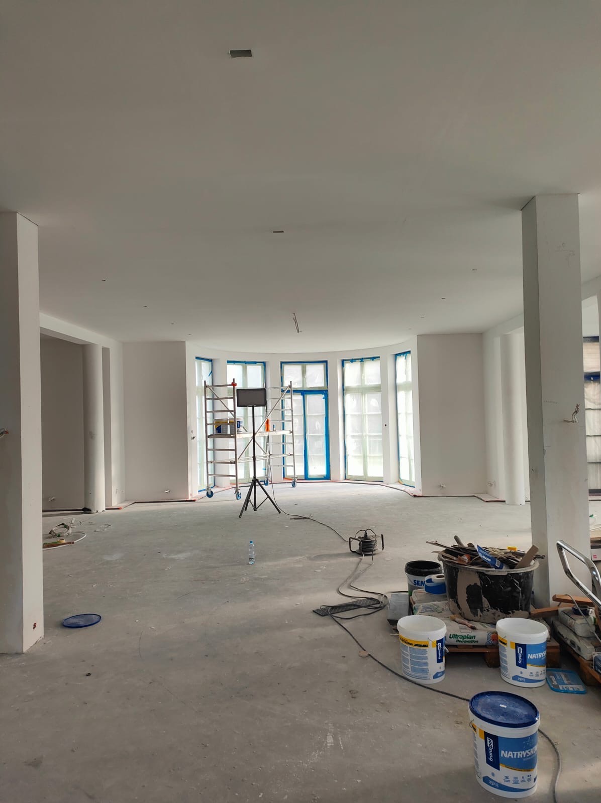 Wnętrze w trakcie prac wykończeniowych, widoczne rusztowanie, okna zabezpieczone folią, wiadra z farbą i narzędzia malarskie na podłodze.