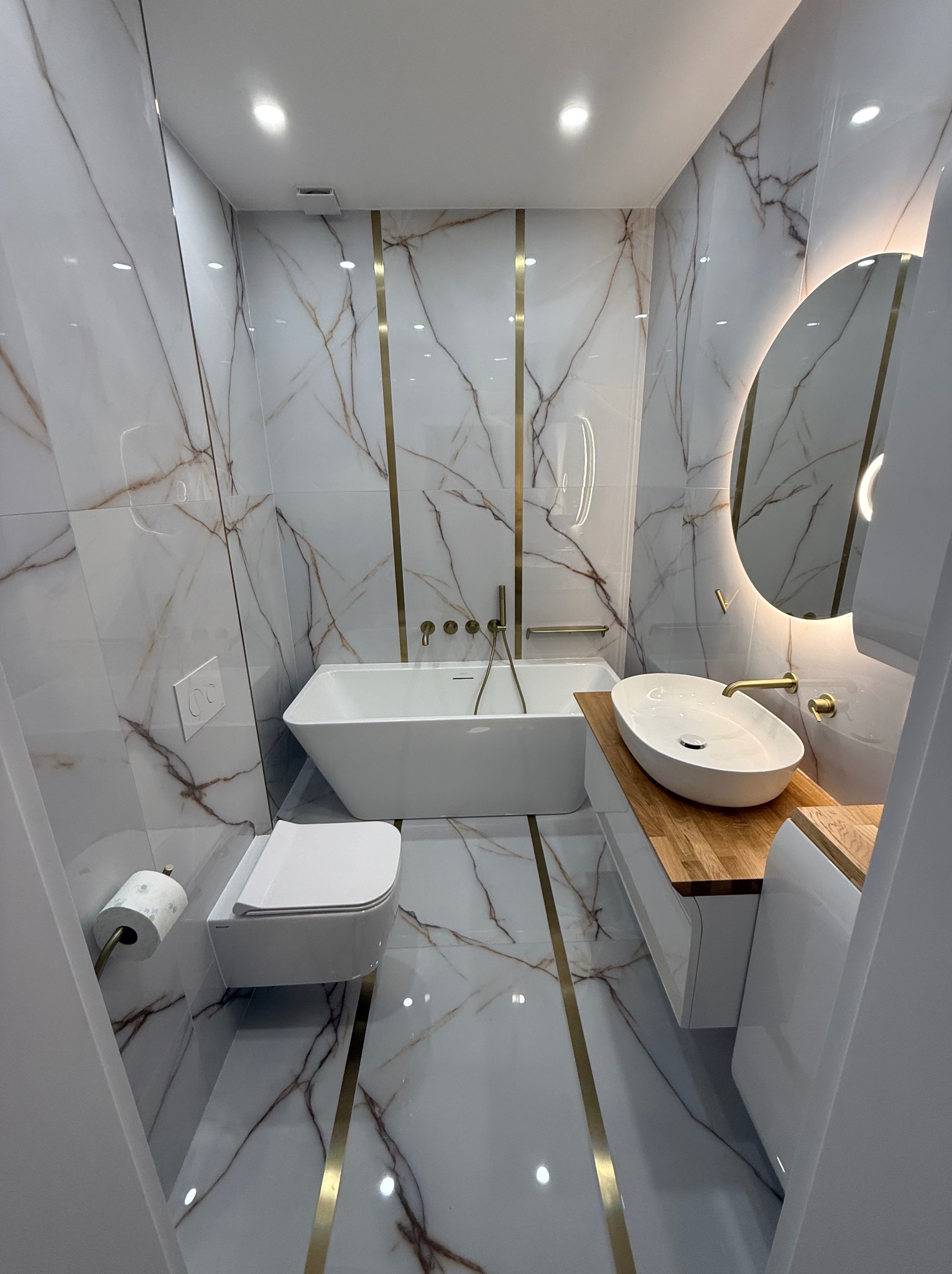 Nowoczesna łazienka z białymi płytkami imitującymi marmur z złotymi akcentami, owalną umywalką na drewnianym blacie, podświetlanym okrągłym lustrem, wiszącą miską WC i wolnostojącą wanną.