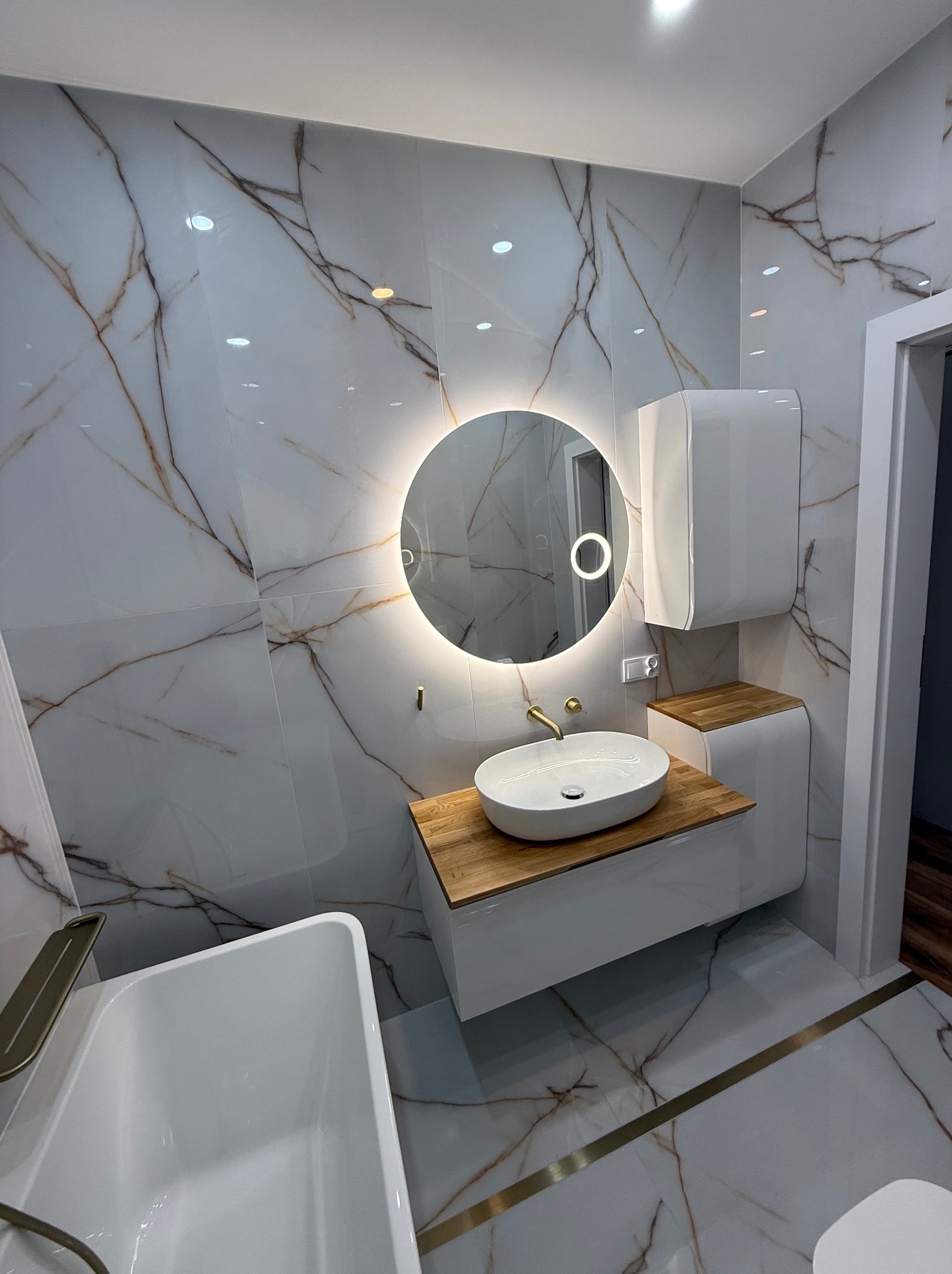 Nowoczesna łazienka z marmurowymi płytkami, okrągłym lustrem z podświetleniem LED, białą umywalką nablatową na drewnianym blacie i białą szafką wiszącą