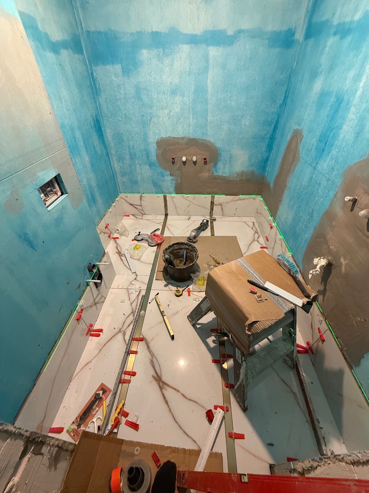 Łazienka w trakcie układania płytek podłogowych z efektem marmuru, ściany przygotowane z hydroizolacją w odcieniach błękitu, widoczne narzędzia i poziomica laserowa.