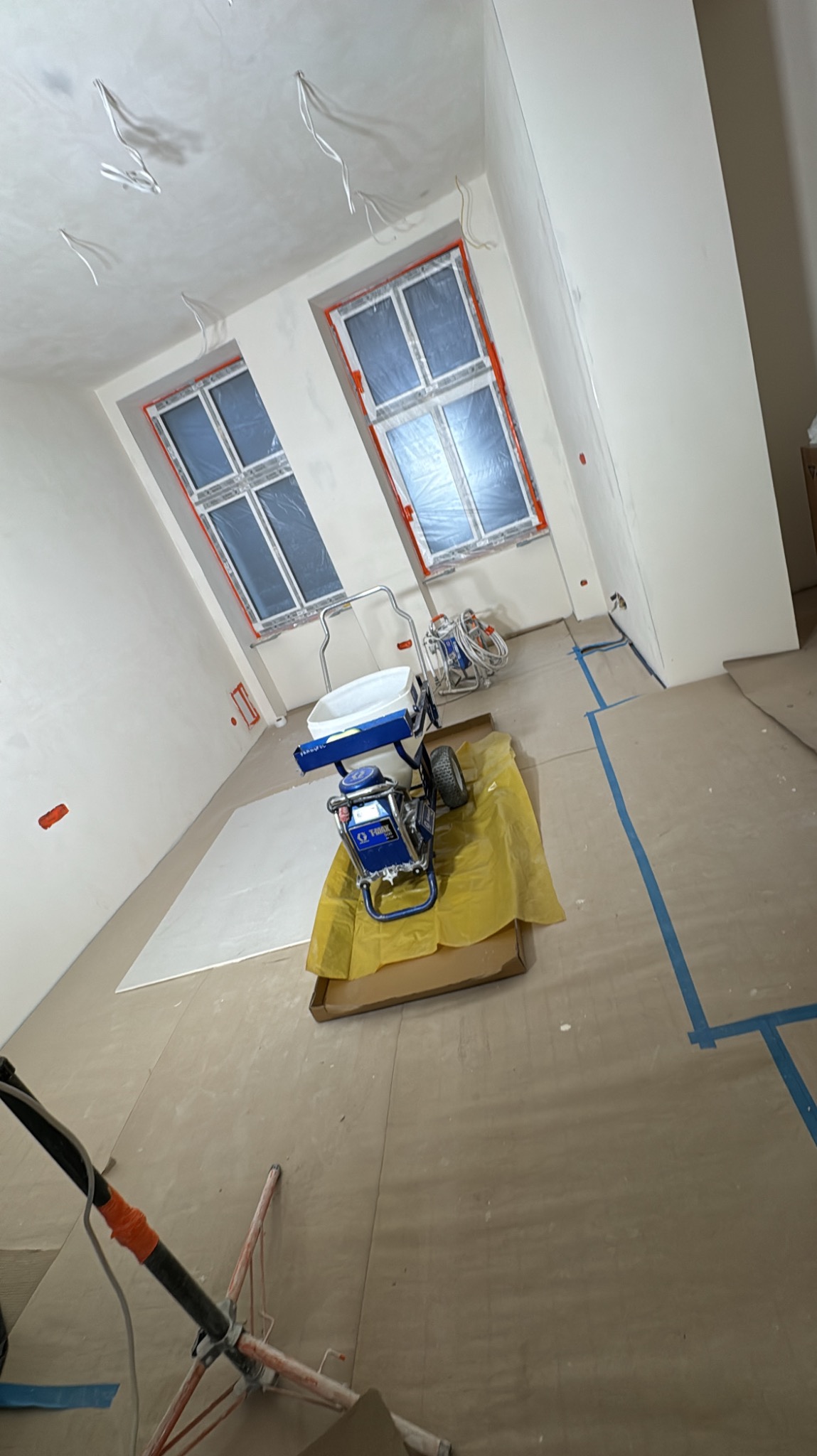 Wnętrze w trakcie prac przygotowawczych do malowania: zaklejone okna folią, zabezpieczona podłoga, widoczny agregat malarski, wystające kable elektryczne z sufitu.