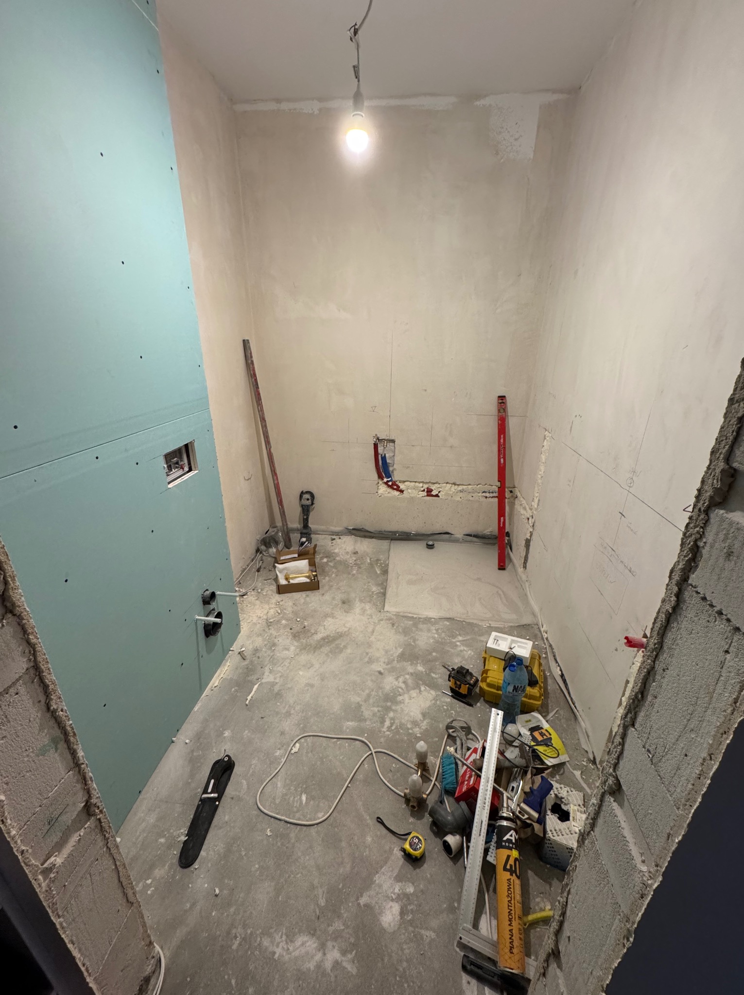 Łazienka w trakcie remontu: ściany z płyt gipsowo-kartonowych, częściowo pokryte gładzią, instalacja wodna i elektryczna widoczna, liczne narzędzia i materiały budowlane na podłodze.