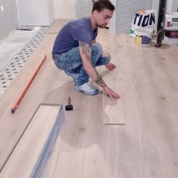 Łukasz Langa - Układanie jasnych paneli podłogowych przez instalatora w dżinsach i crocsach. Widoczny poziomica, młotek i paczki paneli. Remont w trakcie wykańczania.
