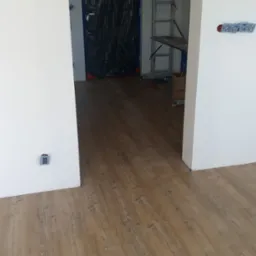 Jasne panele podłogowe z widocznym wzorem drewna w nowym wnętrzu. W tle zabezpieczone drzwi i drabina malarska. Widoczne gniazdka elektryczne.