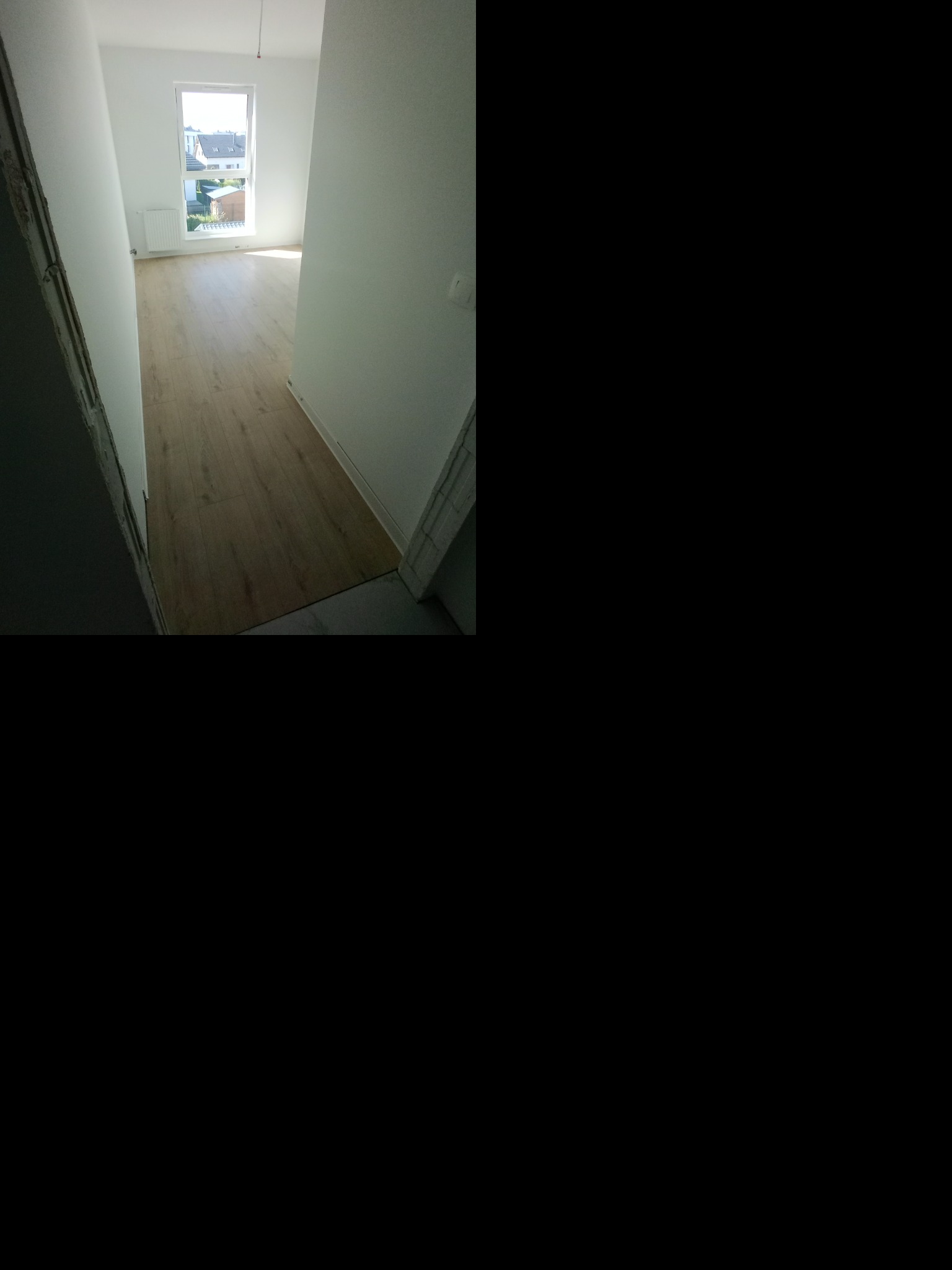 Jasny pokój po remoncie w Wejherowie z panelami podłogowymi i świeżo pomalowanymi ścianami. Widok z korytarza na okno i otoczenie budynku.