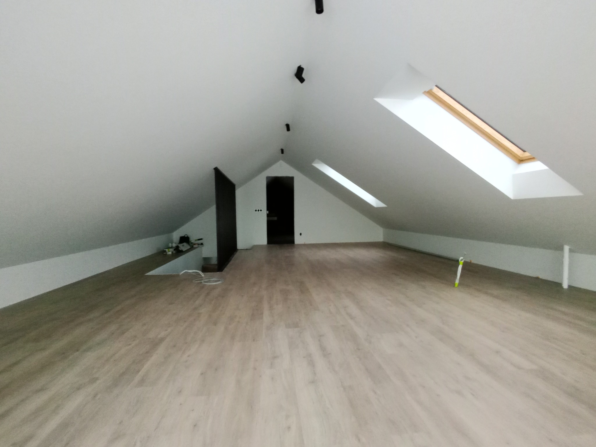 Jasne panele podłogowe na poddaszu z oknami dachowymi i białymi ścianami. Widoczne czarne lampy sufitowe i częściowo wyposażenie. Minimalistyczny i nowoczesny design.