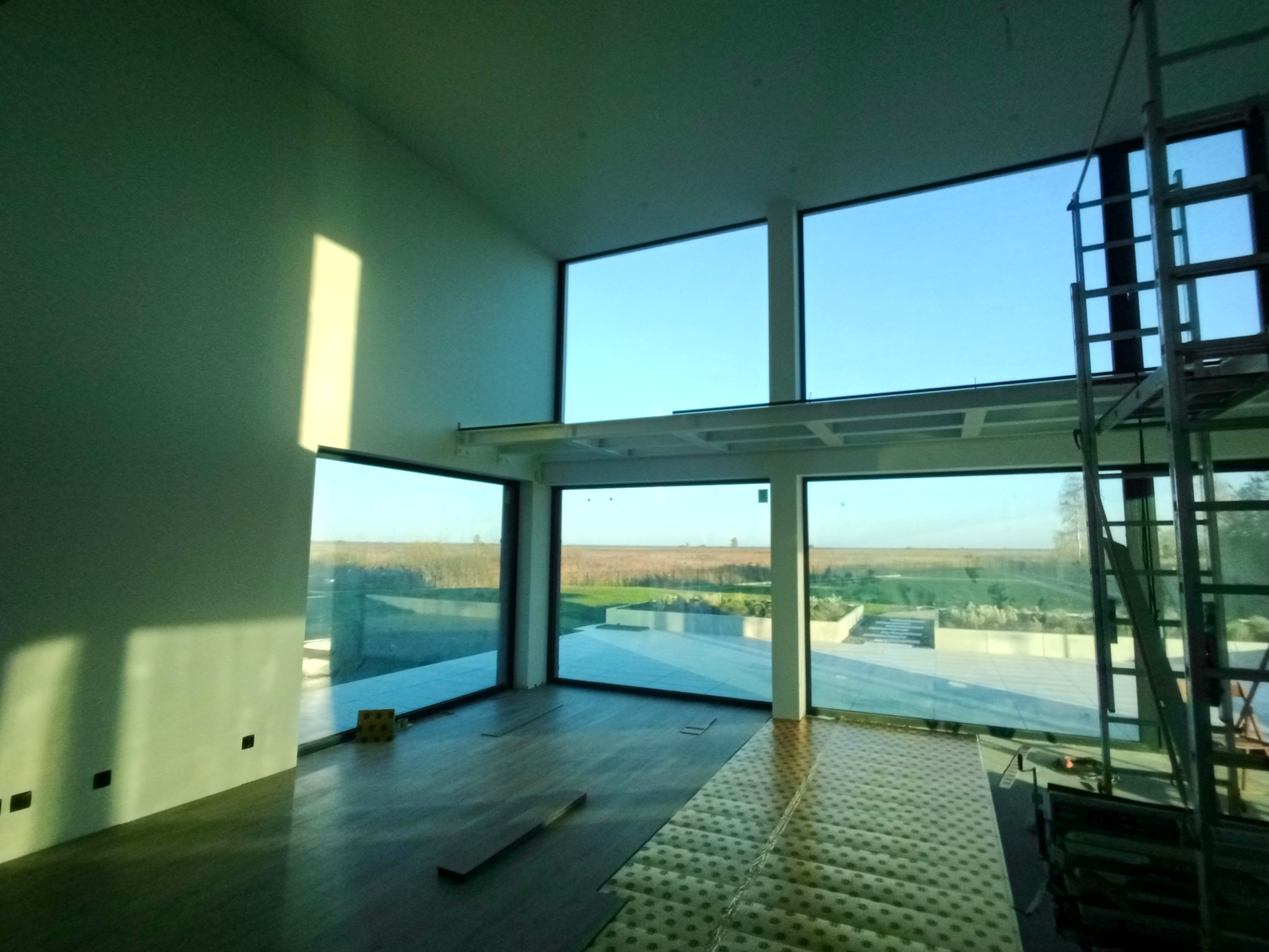 Wnętrze z dużymi oknami i częściowo ułożonymi panelami podłogowymi. Widoczny krajobraz za oknem i drabina malarska. Ściana oświetlona popołudniowym słońcem.