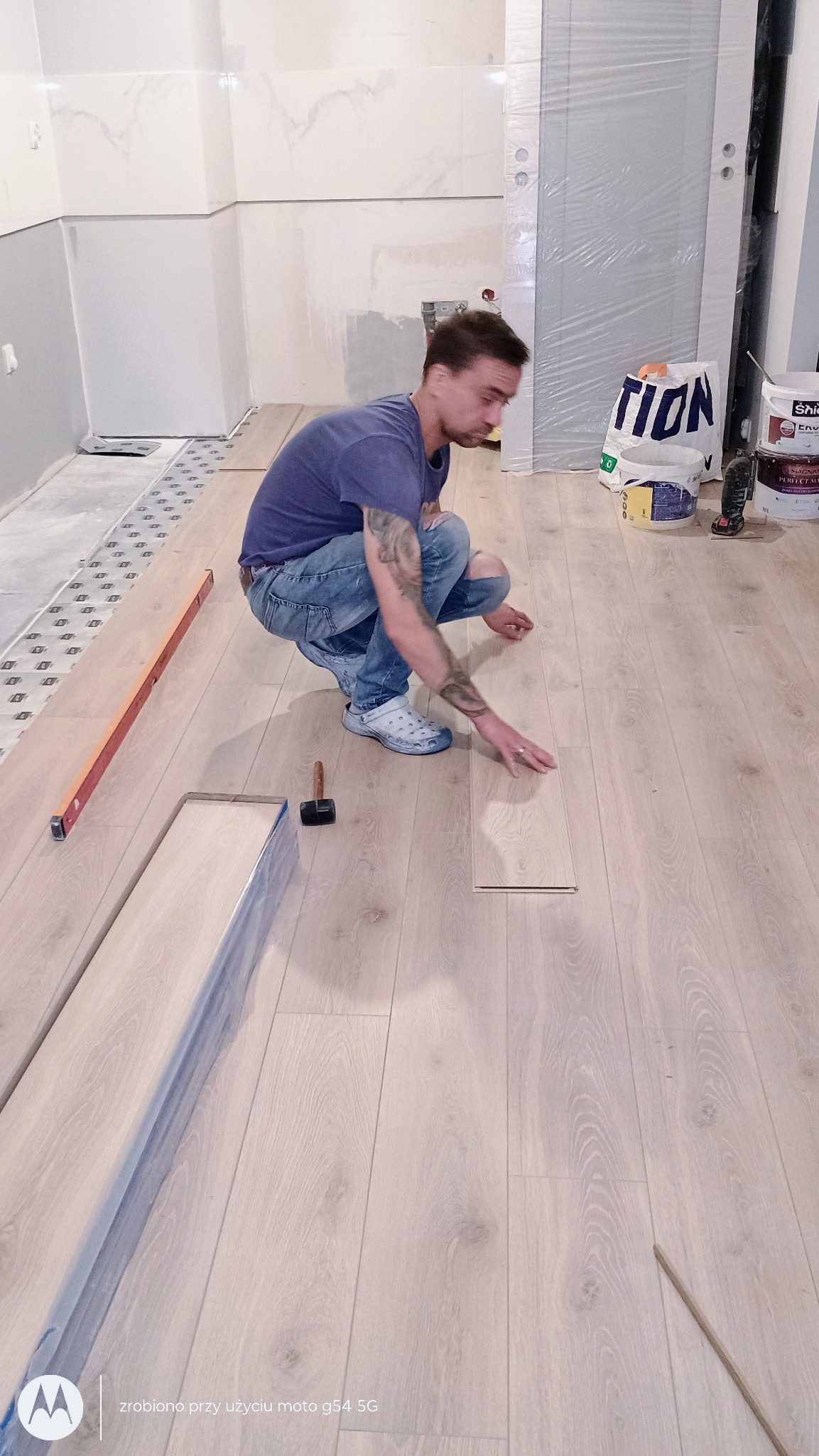 Układanie jasnych paneli podłogowych przez instalatora w dżinsach i crocsach. Widoczny poziomica, młotek i paczki paneli. Remont w trakcie wykańczania.