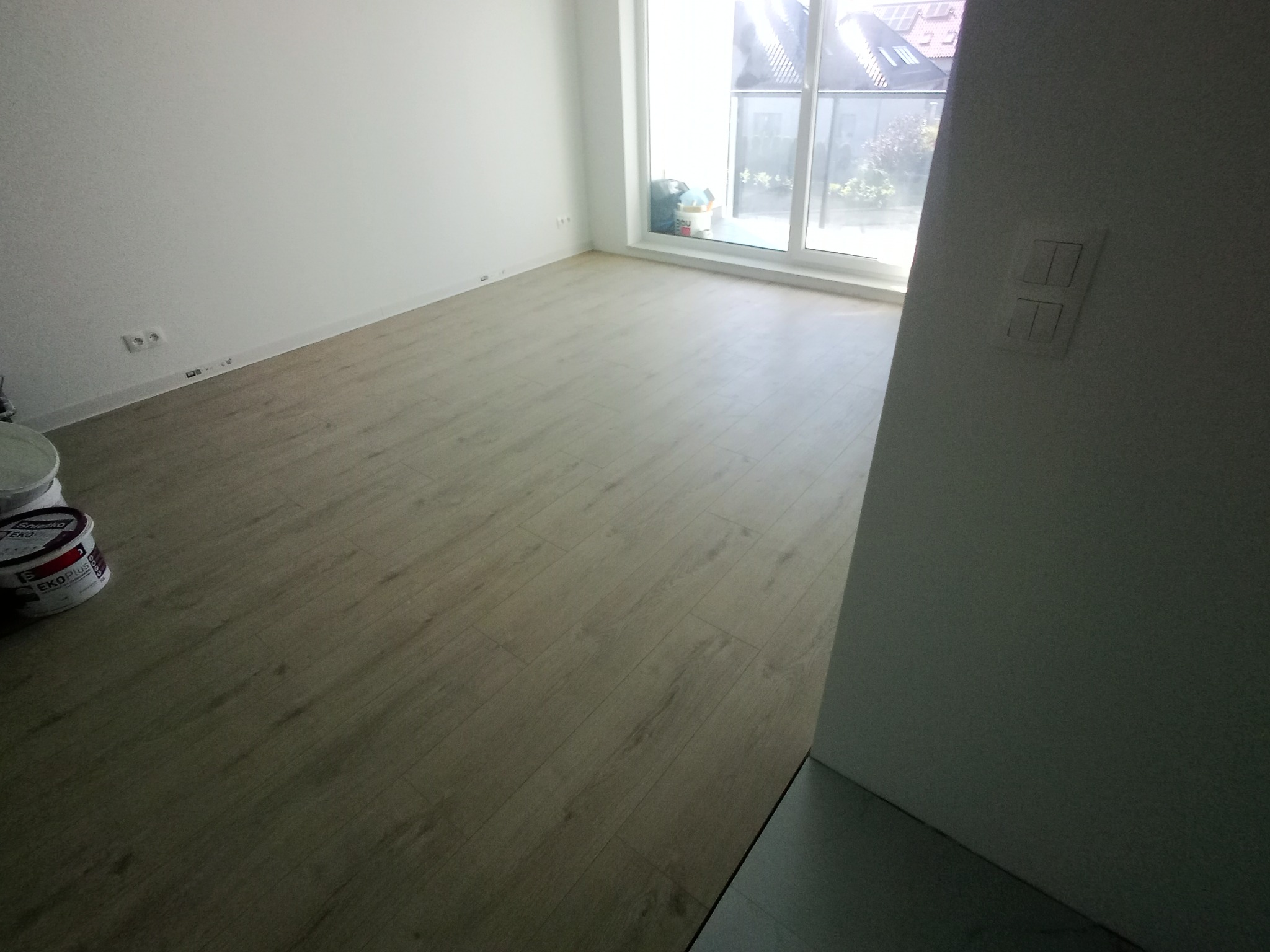 Jasne panele podłogowe w pokoju z białymi ścianami i dużym oknem. Widoczne puszki z farbą EKO PLUS przy ścianie. Minimalistyczne wnętrze po remoncie.