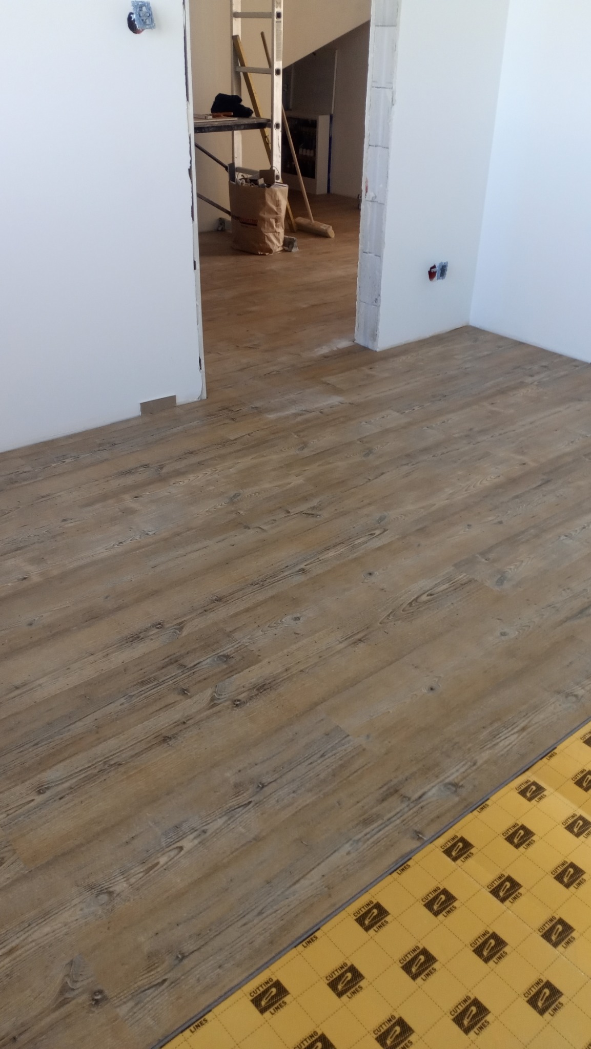 Jasnobrązowe panele podłogowe imitujące drewno, częściowo ułożone, z widoczną podkładką izolacyjną. W tle ściana i drabina w przejściu do innego pomieszczenia.