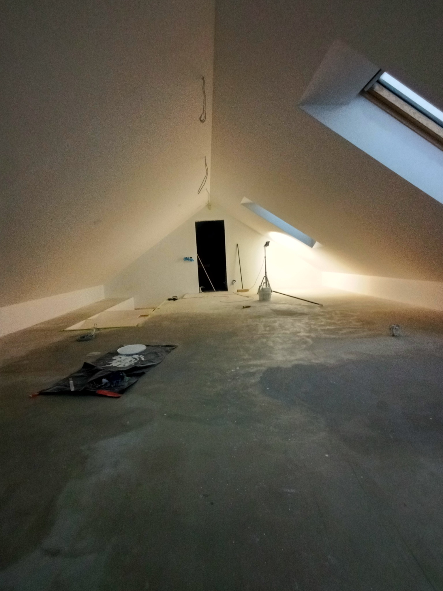 Poddasze w trakcie prac wykończeniowych: szpachlowanie ścian, widoczne okna dachowe, lampa budowlana, narzędzia malarskie na podłodze. Ściany przygotowane do malowania.