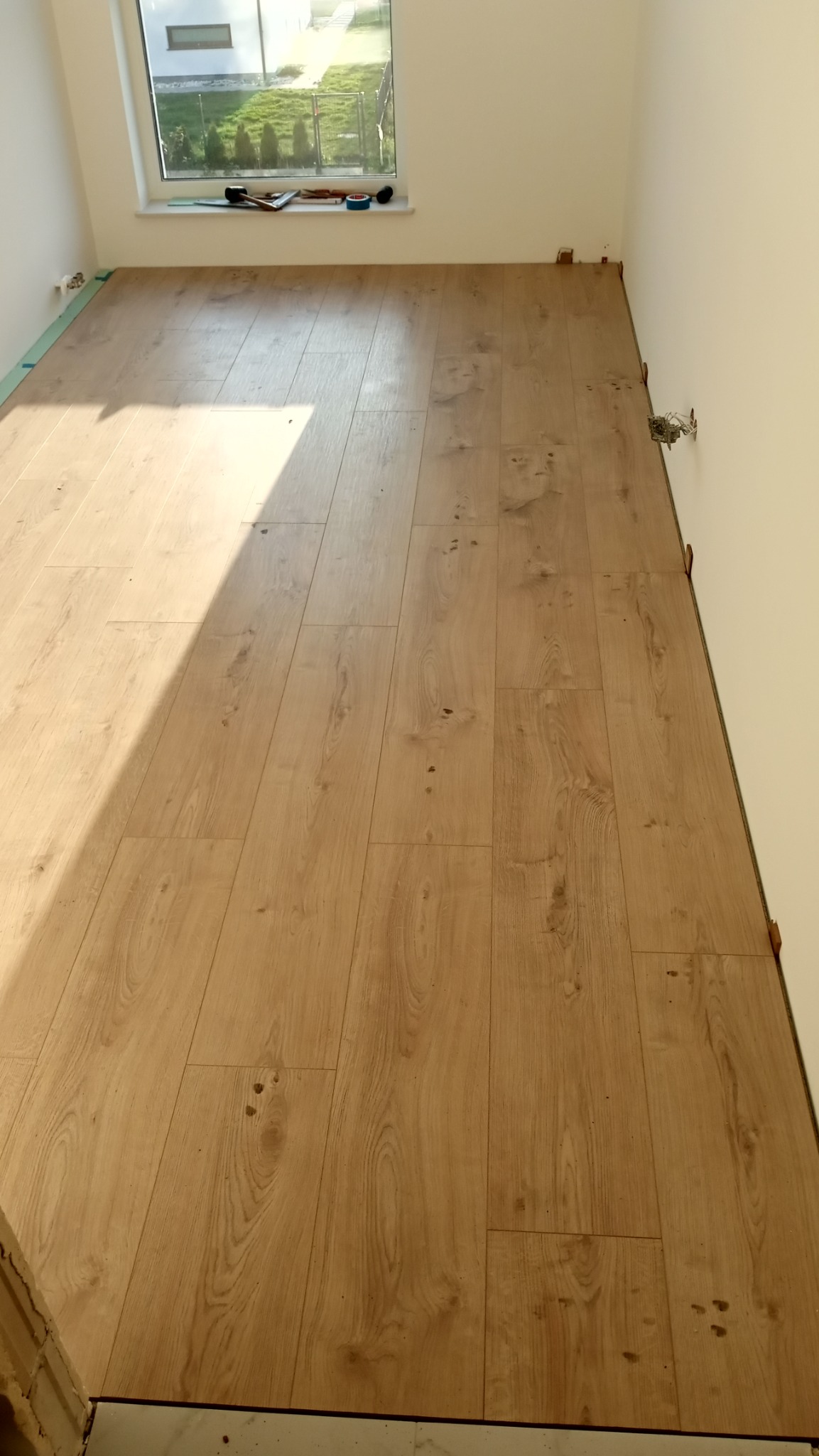 Jasne panele podłogowe ułożone w pokoju z oknem, widoczne narzędzia na parapecie. Częściowo wykończone, z widocznymi elementami mocującymi przy ścianie.