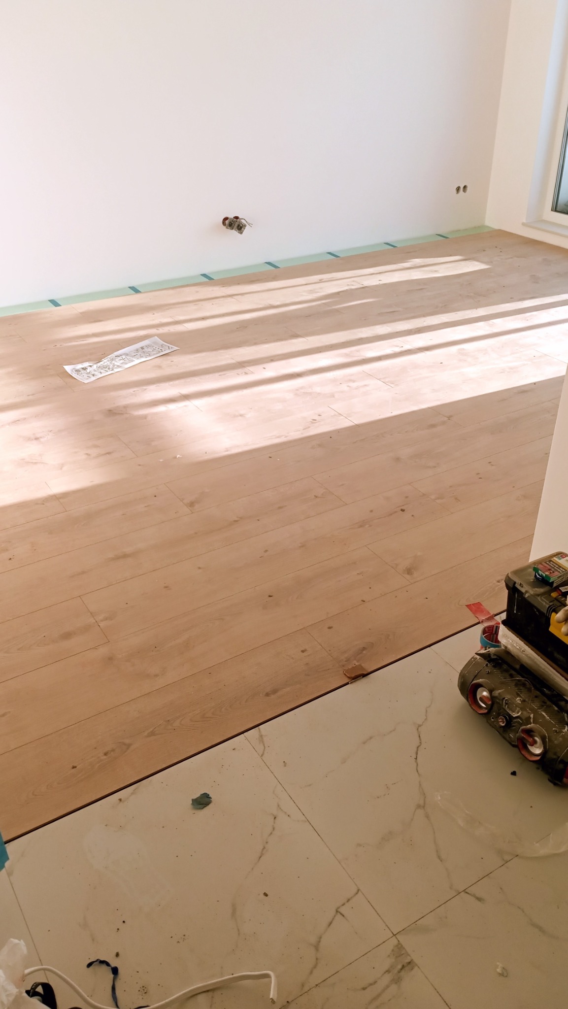 Jasne panele podłogowe w trakcie montażu, widoczne narzędzia i taśma zabezpieczająca ściany. Częściowo ułożona podłoga w słonecznym pokoju.