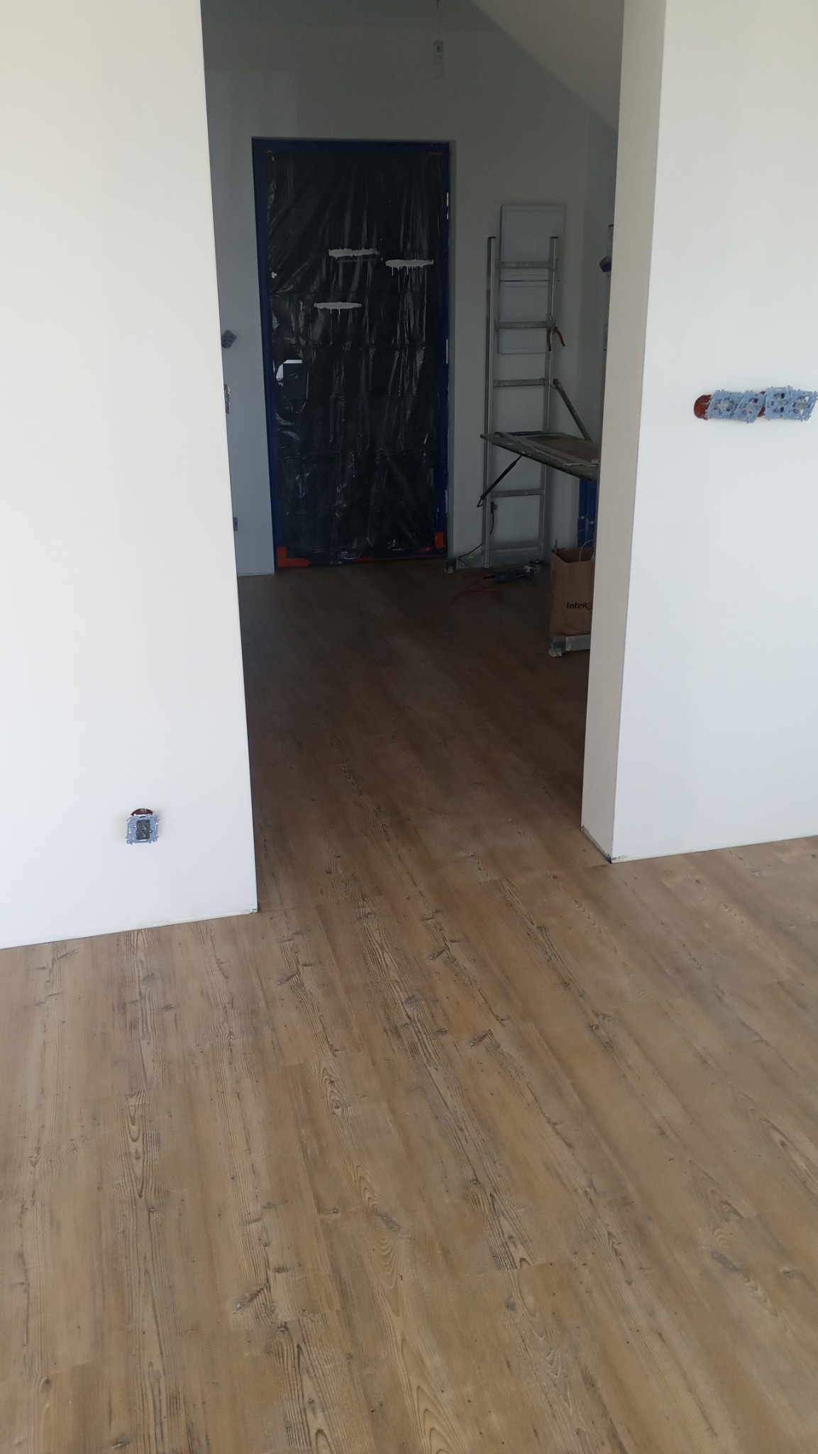 Jasne panele podłogowe z widocznym wzorem drewna w nowym wnętrzu. W tle zabezpieczone drzwi i drabina malarska. Widoczne gniazdka elektryczne.