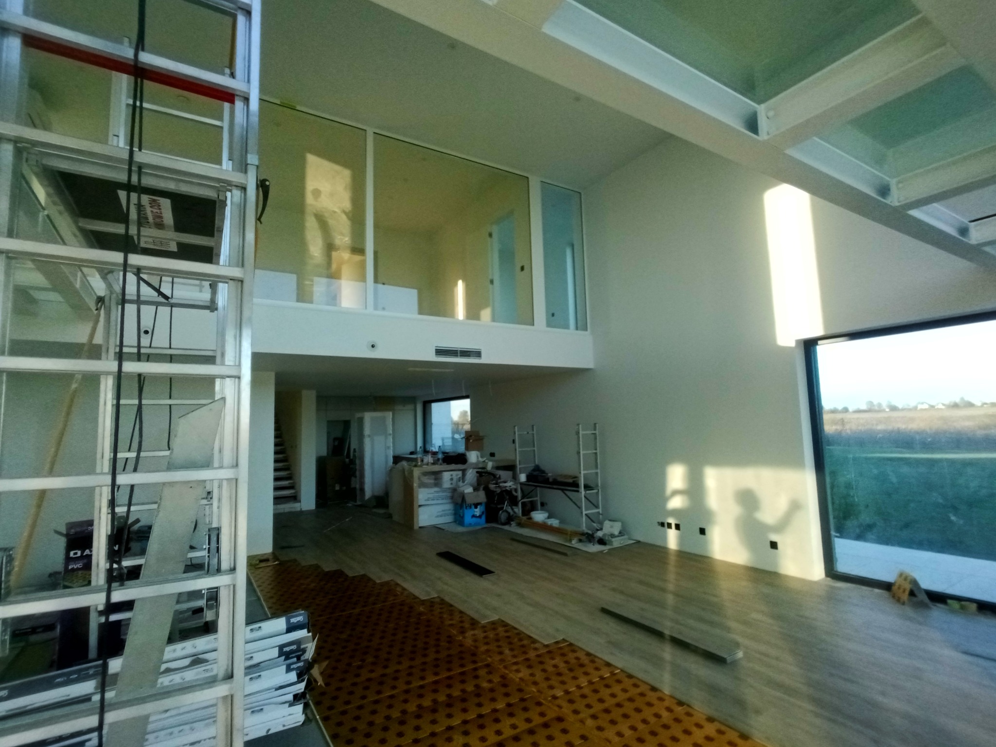 Wnętrze domu w trakcie układania paneli podłogowych, widoczna drabina i okna. Część paneli ułożona na podkładzie. Duże okno z widokiem na zewnątrz.