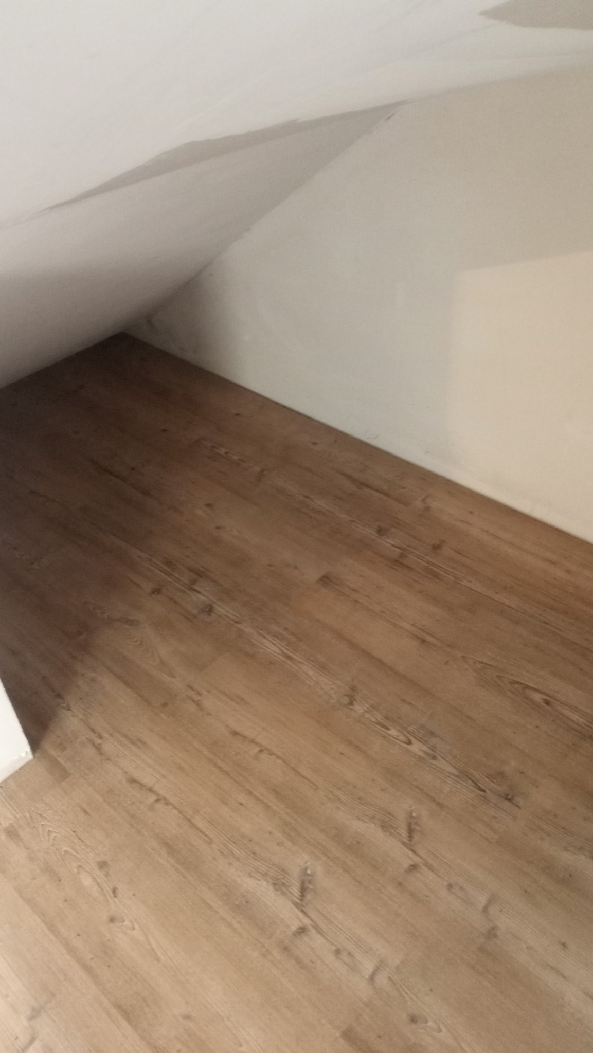 Ukończone układanie paneli podłogowych w pomieszczeniu ze skosem, widoczne panele o jasnym, drewnopodobnym wzorze, przy białej ścianie. Ujęcie z góry.