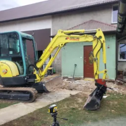 Żółto-czarna mini koparka Ammann Yanmar na gąsienicach, z łyżką i lemieszem, stoi na trawie przed budynkiem. Widoczny laser krzyżowy na statywie. Rozpoczęte prace ziemne.