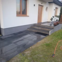 Ułożona, ciemnoszara kostka brukowa prowadząca do wejścia domu z trzema schodami, widoczne donice z roślinami ozdobnymi, trawnik i wąż ogrodowy.