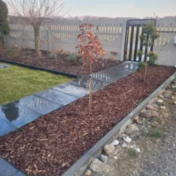 Ścieżka z ciemnych płyt prowadząca do furtki, obłożona korą, obok trawnik i fragment ogrodzenia z automatyką