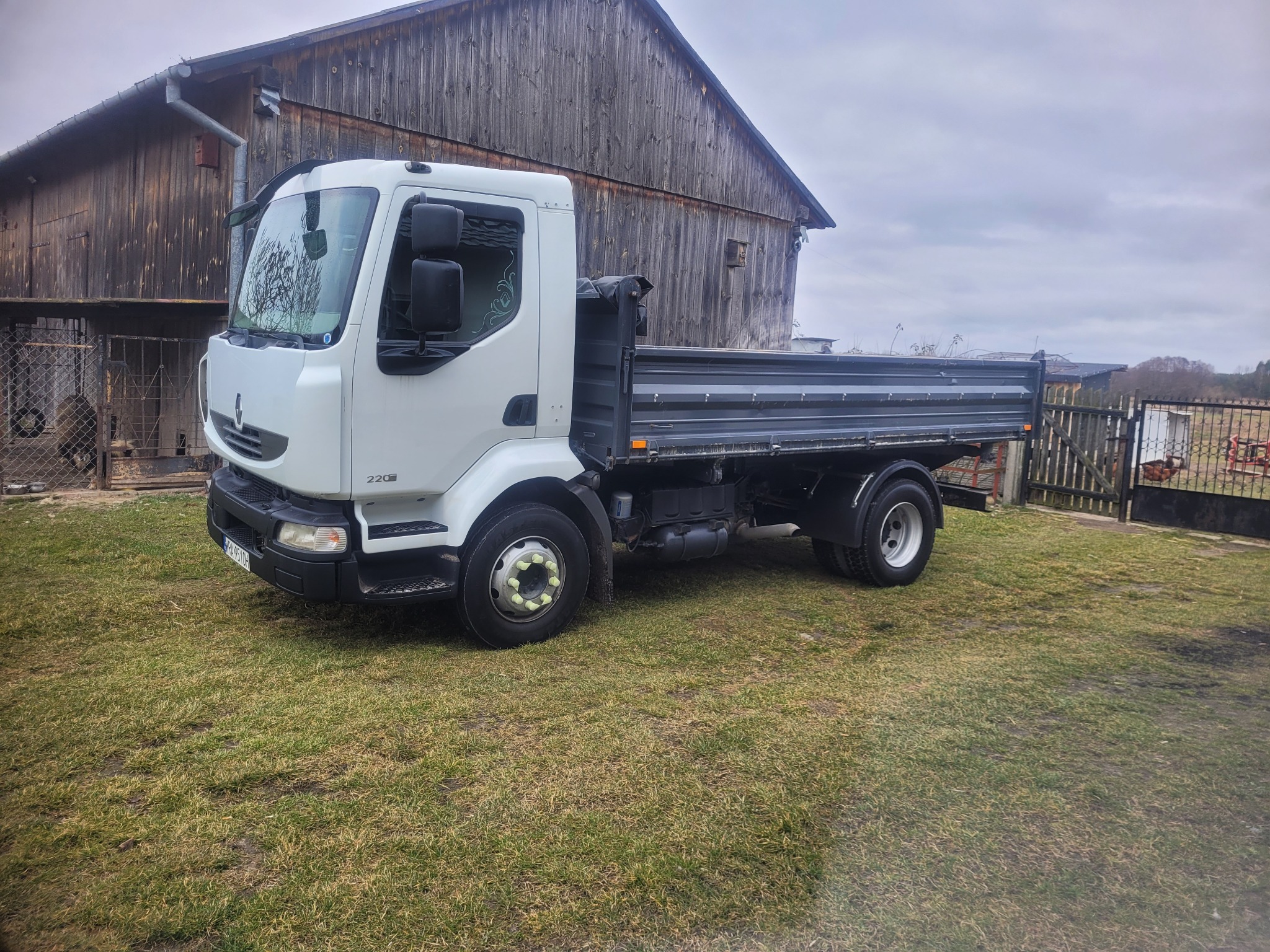 Biała wywrotka Renault na trawiastym terenie przed drewnianym budynkiem gospodarczym w pochmurny dzień.