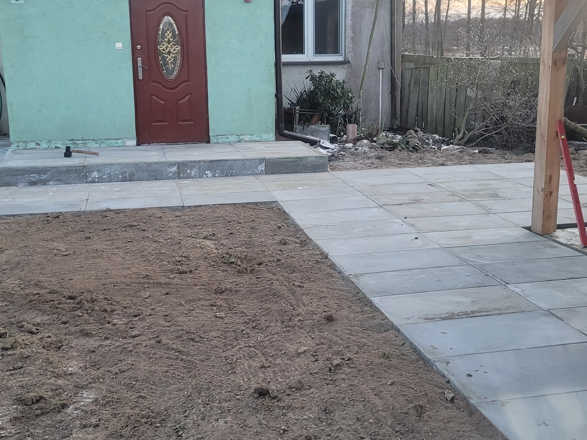 Układanie szarej kostki brukowej na posesji, widoczny młotek na schodach, częściowo przygotowane podłoże z ziemi, poziomica oparta o drewniany słup.