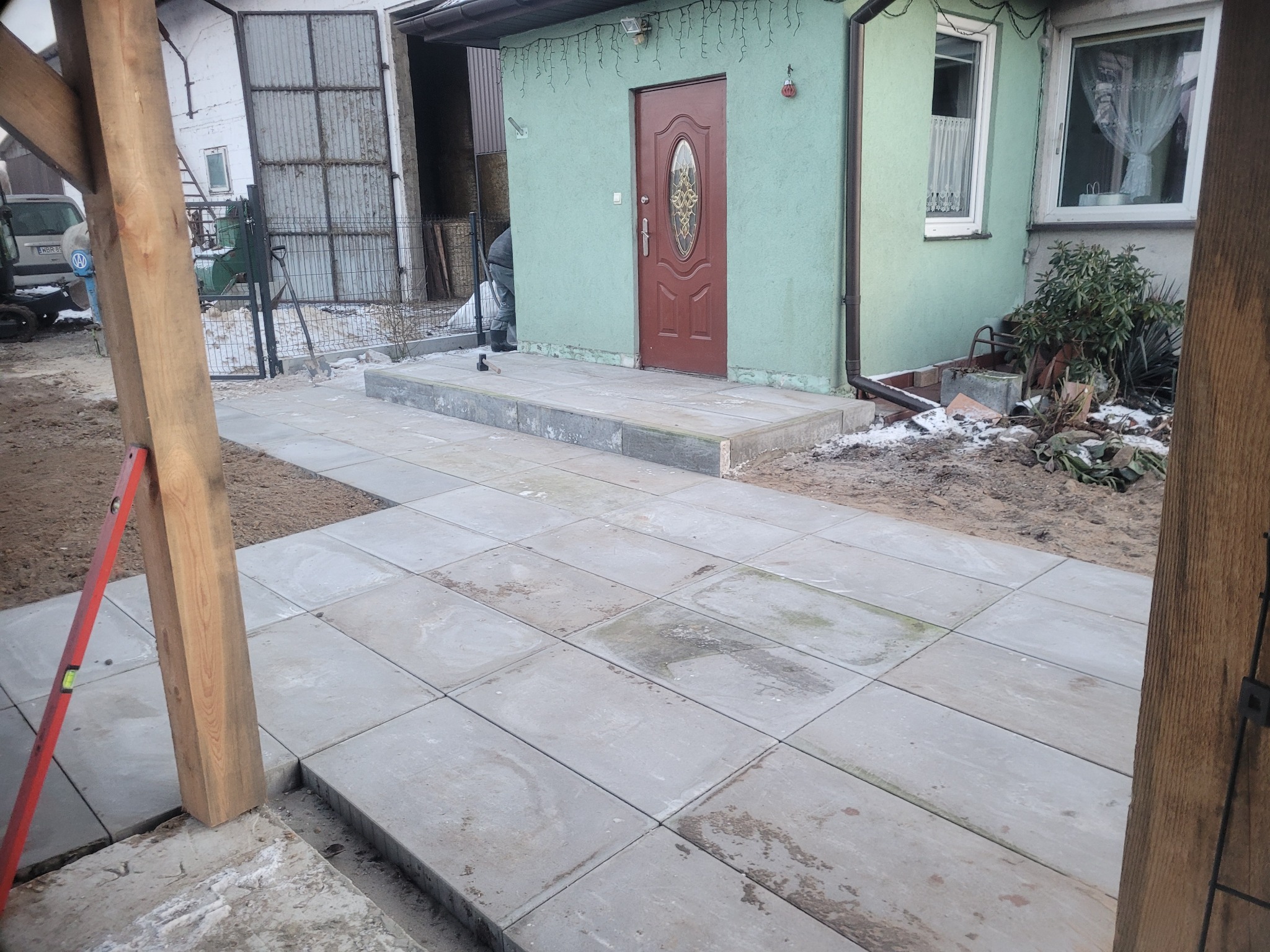 Ułożona szara kostka brukowa prowadząca do wejścia domu z zieloną elewacją i brązowymi drzwiami, widoczny poziomica oparta o drewniany słup.