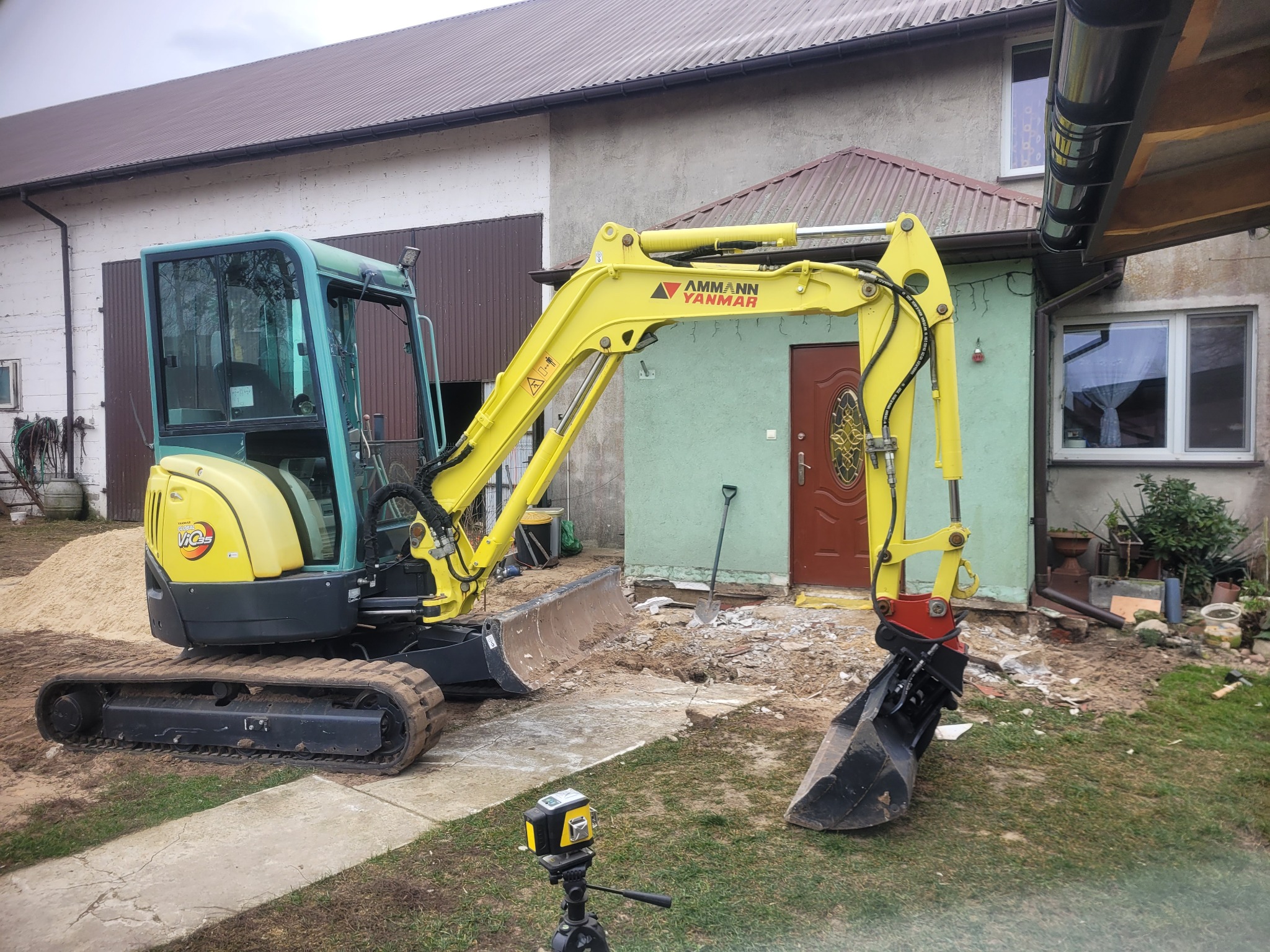 Żółto-czarna mini koparka Ammann Yanmar na gąsienicach, z łyżką i lemieszem, stoi na trawie przed budynkiem. Widoczny laser krzyżowy na statywie. Rozpoczęte prace ziemne.