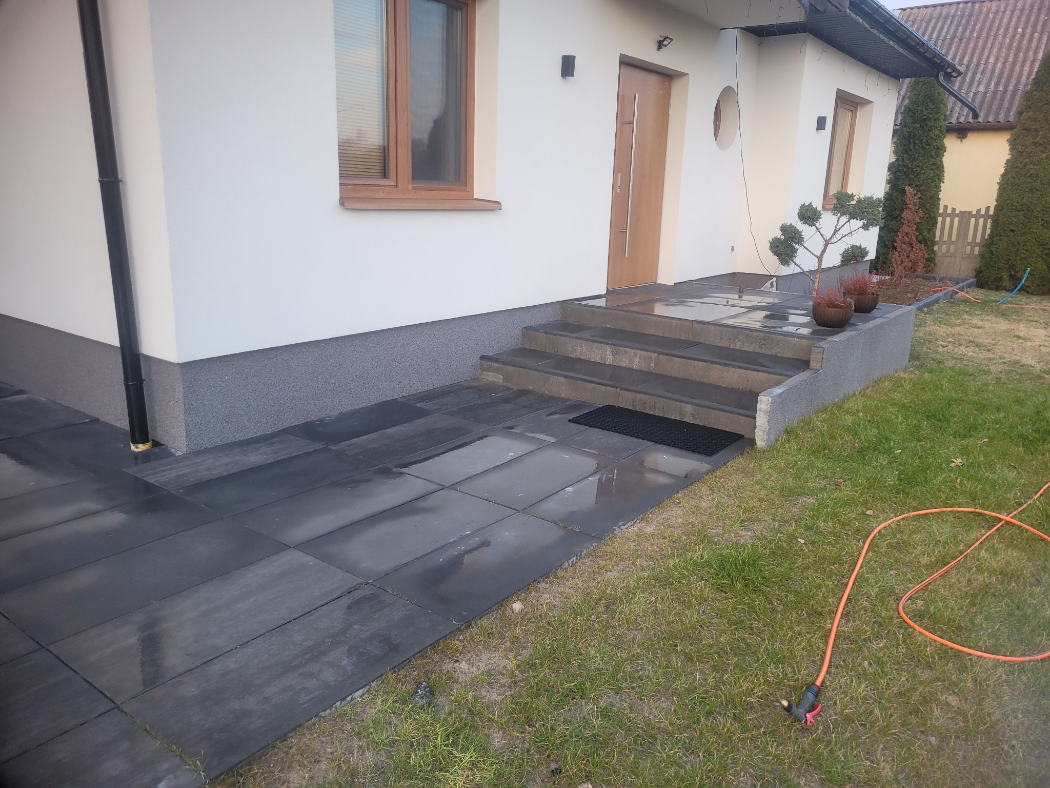 Ułożona, ciemnoszara kostka brukowa prowadząca do wejścia domu z trzema schodami, widoczne donice z roślinami ozdobnymi, trawnik i wąż ogrodowy.