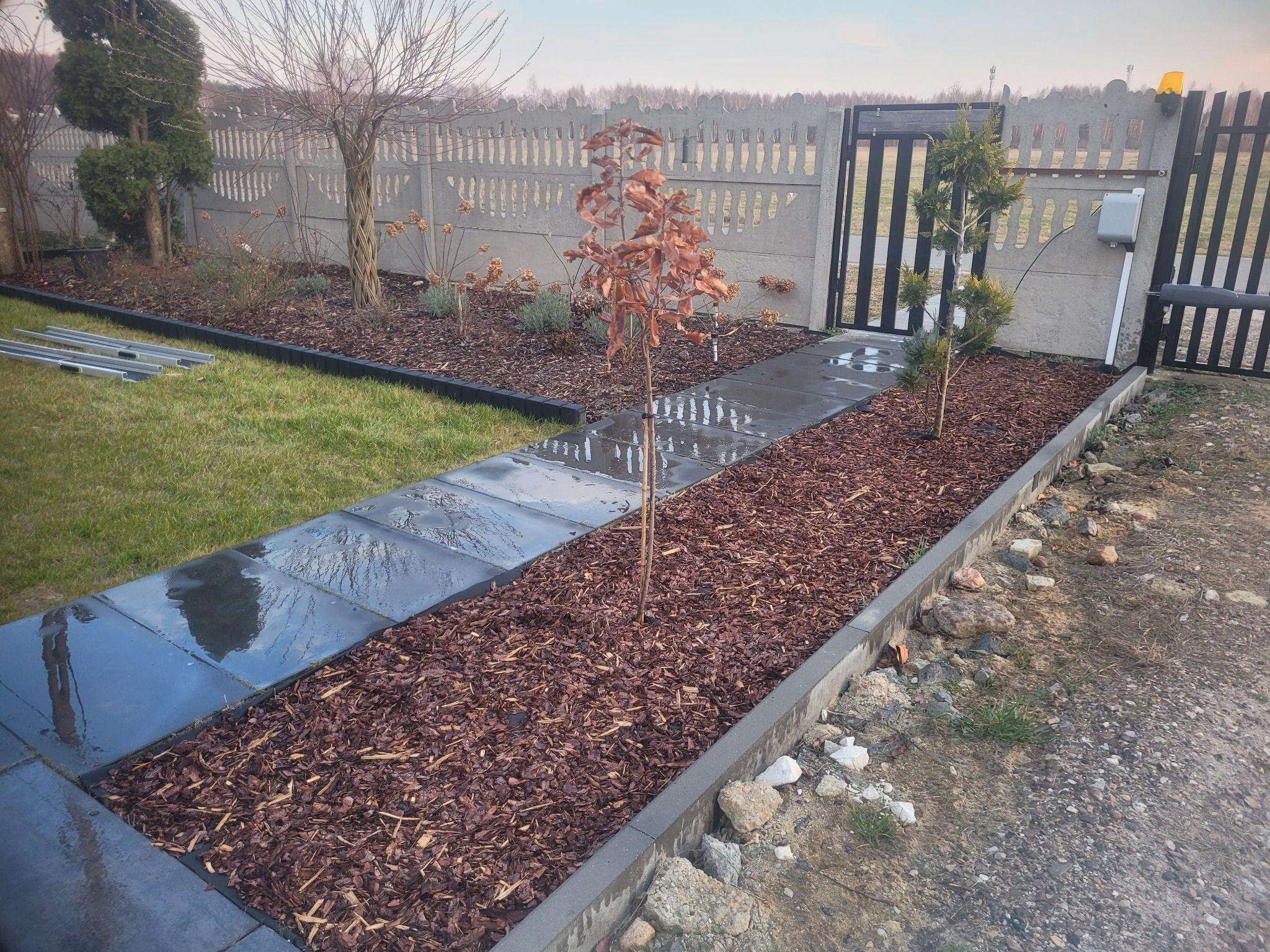 Ścieżka z ciemnych płyt prowadząca do furtki, obłożona korą, obok trawnik i fragment ogrodzenia z automatyką