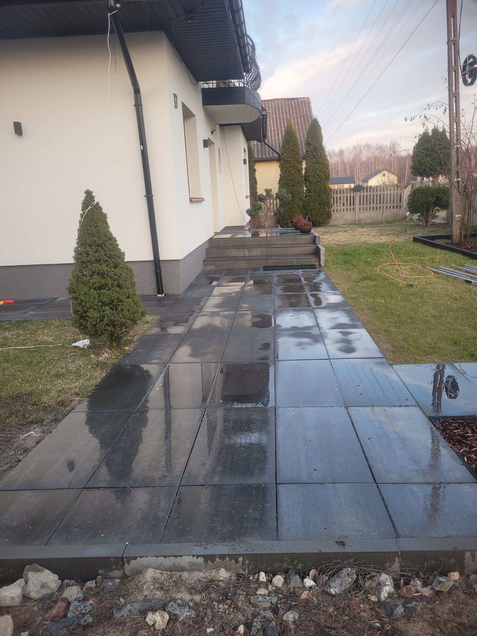 Świeżo ułożony, mokry chodnik z szarej kostki brukowej prowadzący do schodów wejściowych domu jednorodzinnego. Widoczne elementy ogrodu, trawnik i iglaki.