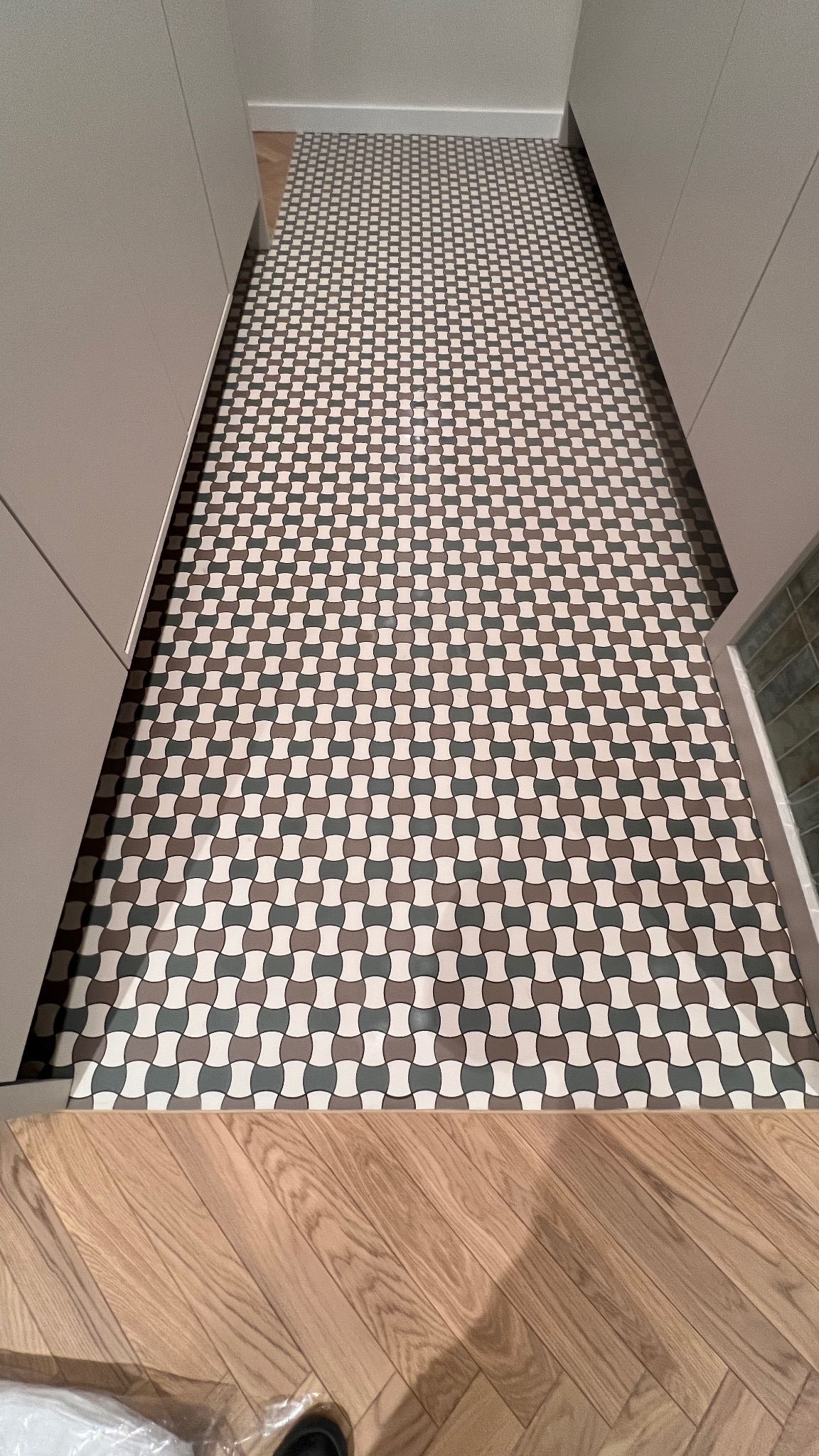 Wzorzysta podłoga z płytek w odcieniach szarości, brązu i bieli, ułożona pomiędzy jasnymi szafkami, przechodząca w drewnianą podłogę w przedpokoju.