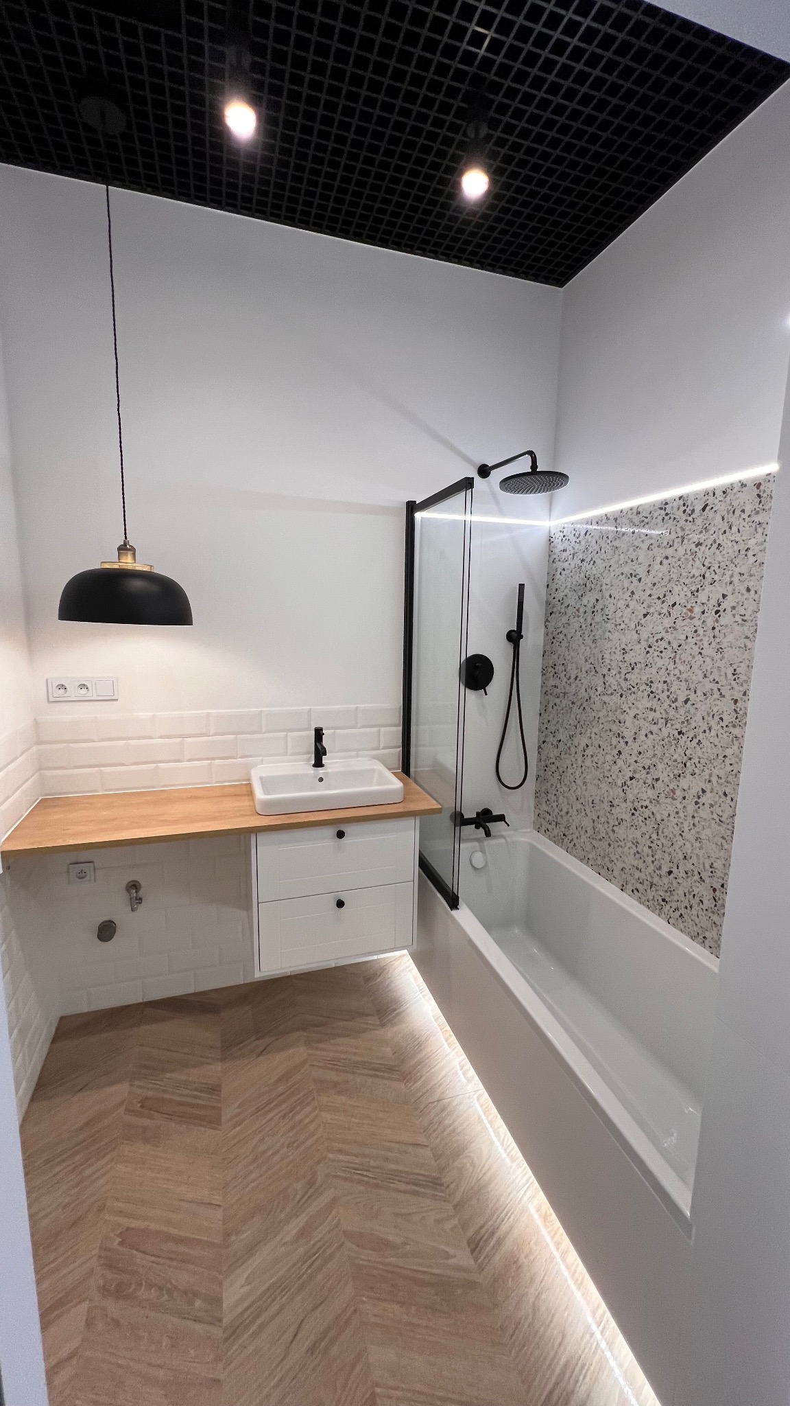 Nowoczesna łazienka z białą armaturą, drewnianym blatem, czarną baterią i kabiną prysznicową z czarnymi profilami, podświetleniem LED i wzorzystą ścianą.