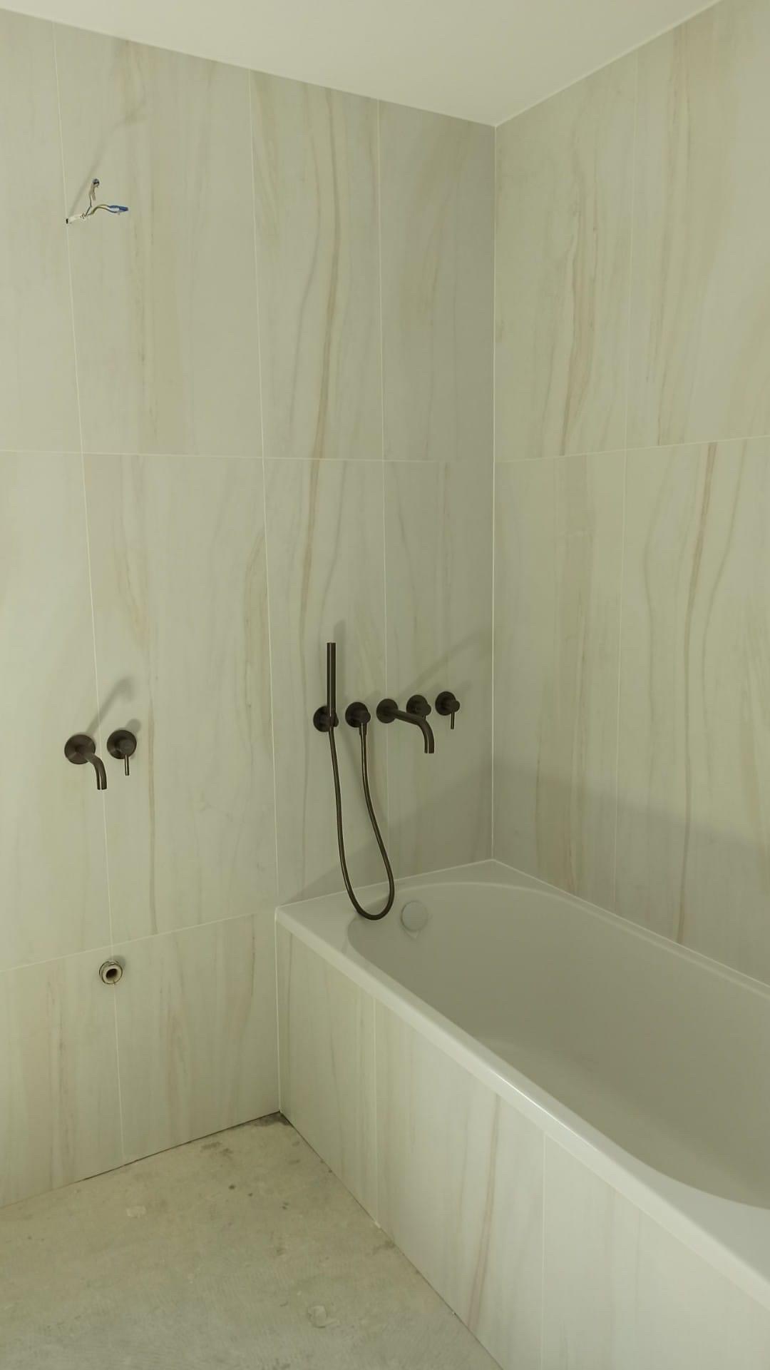 Ściana łazienki wyłożona płytkami imitującymi marmur, z zamontowaną wanną i baterią w kolorze ciemnego brązu, widoczne niedokończone podłączenie elektryczne.