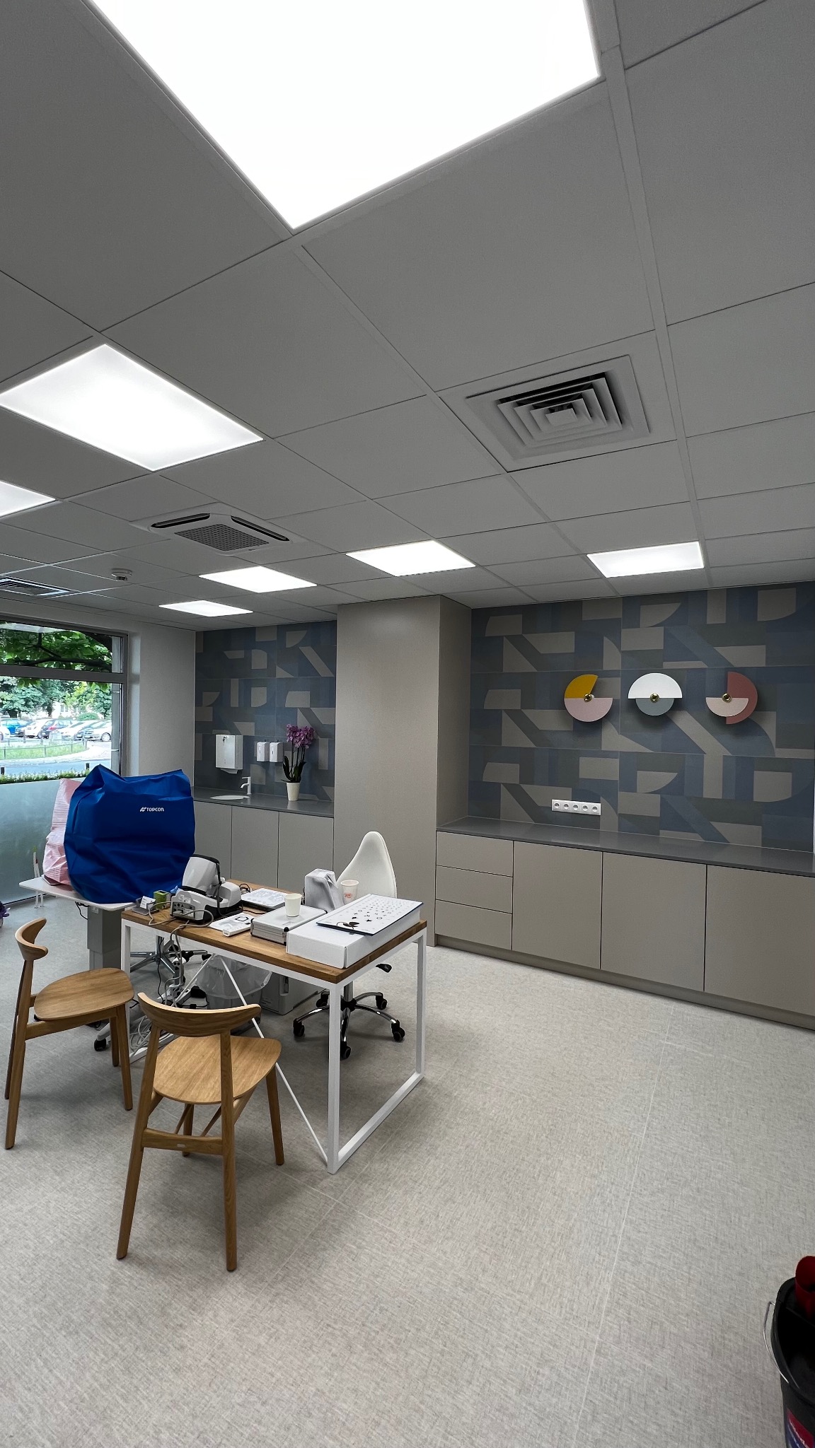 Wnętrze biura z geometrycznym wzorem na ścianie, jasne meble, drewniane krzesła i nowoczesne oświetlenie sufitowe.