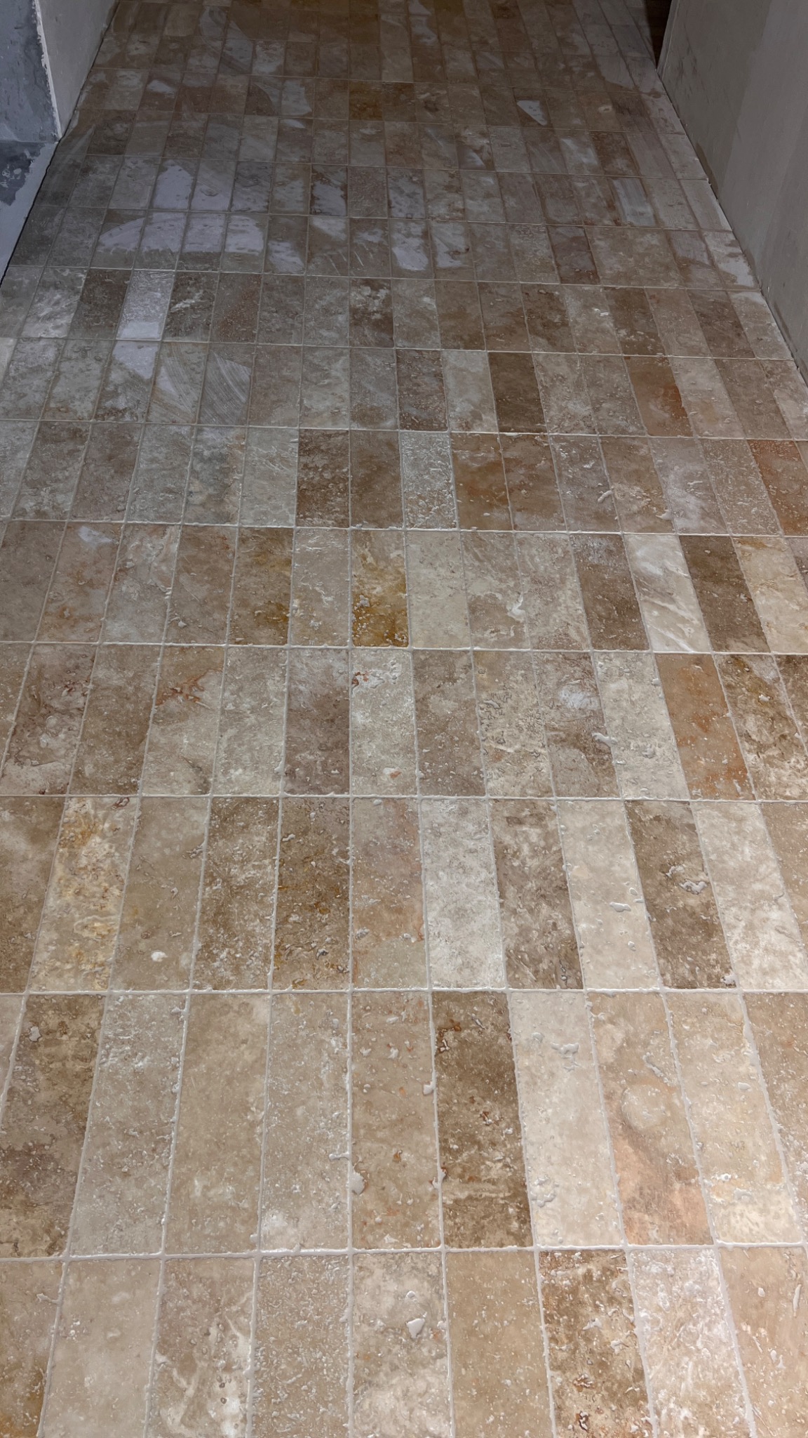 Ułożona, beżowo-brązowa podłoga z płytek w kształcie prostokątów, z widocznymi fugami. Płytki o nieregularnym wzorze imitującym kamień naturalny.