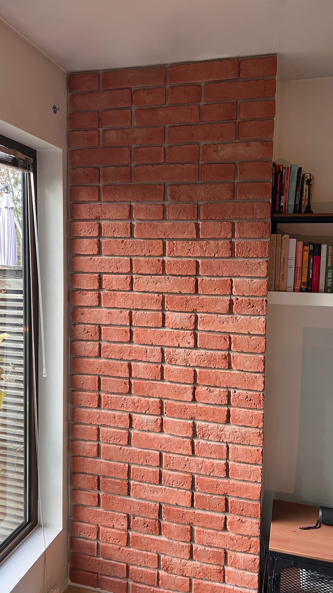 Ściana z czerwonej cegły dekoracyjnej, przylegająca do okna i półki z książkami. Widoczne detale faktury cegieł i spoin. Oświetlenie naturalne, wnętrze.