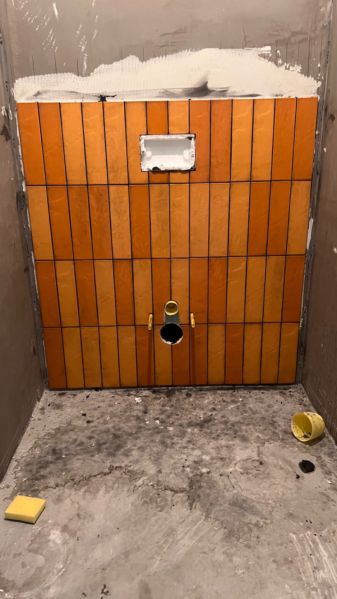 Pomarańczowe, pionowe płytki w trakcie układania na ścianie łazienki, widoczny otwór na instalację i puszka elektryczna. Na podłodze gąbka i wiaderko.