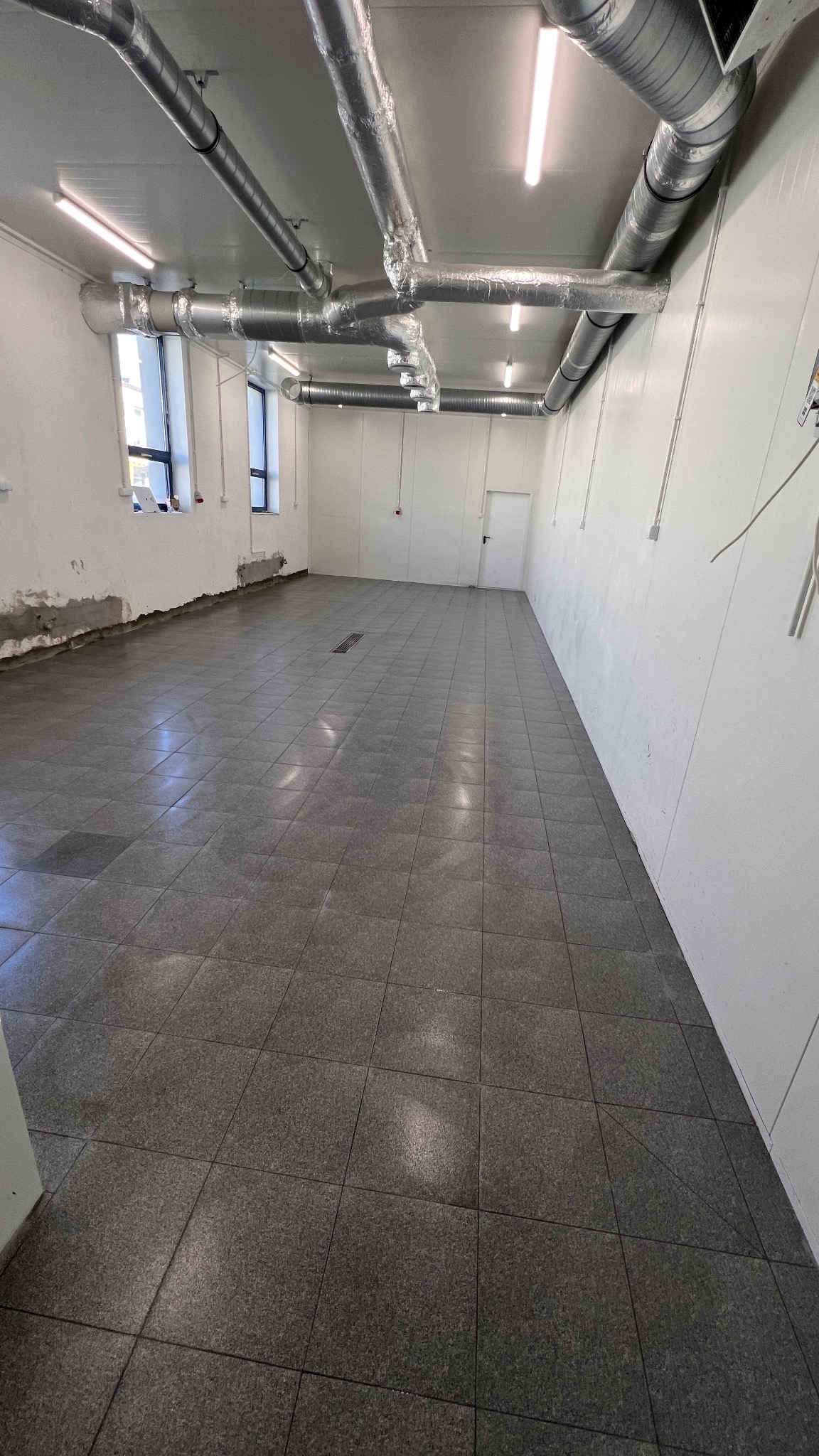 Wykończone wnętrze hali z szarą, kwadratową podłogą z płytek. Widoczne rury wentylacyjne pod sufitem i białe ściany. Fragment ściany z widocznymi śladami po pracach.