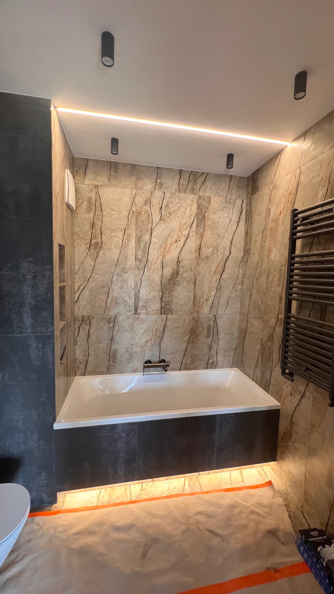 Nowoczesna łazienka z wanną obudowaną ciemnymi płytkami, podświetleniem LED i marmurowymi płytkami na ścianach. Widoczny grzejnik drabinkowy i punktowe oświetlenie sufitowe.