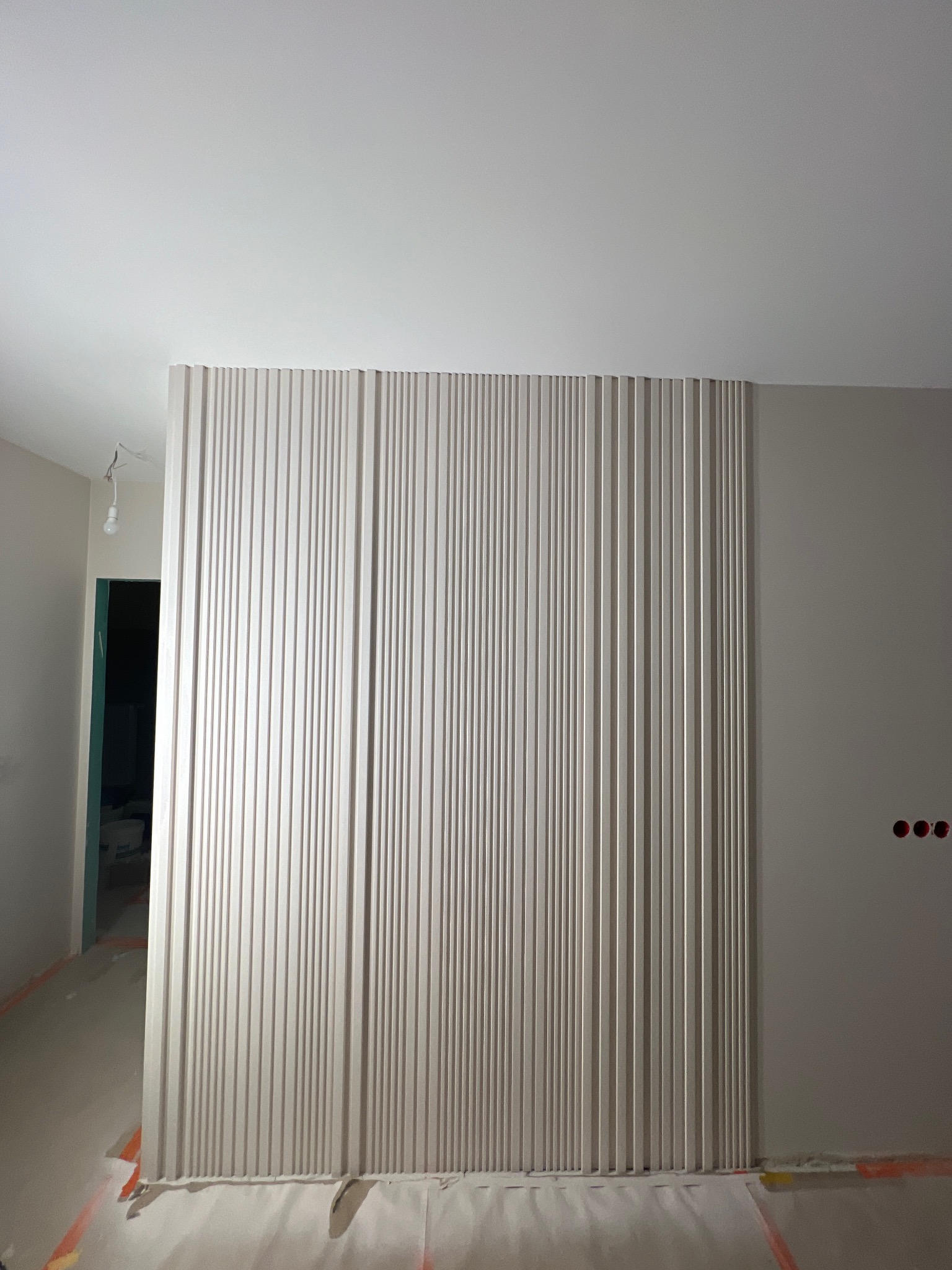 Panel dekoracyjny z pionowych listew w jasnym odcieniu, zamontowany na ścianie w trakcie prac wykończeniowych. Widoczne zabezpieczenia podłogi.