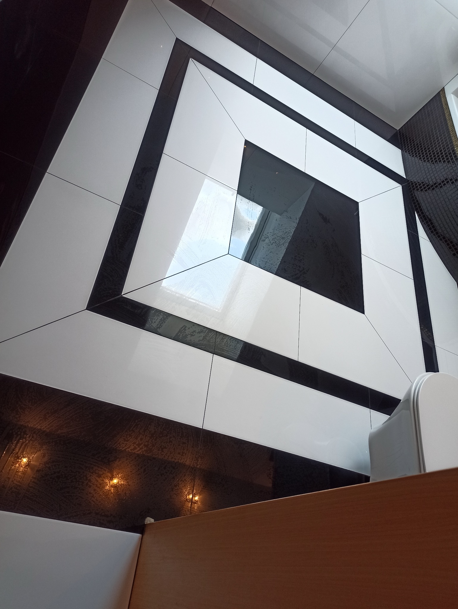 Geometryczny wzór z czarno-białych płytek na podłodze łazienki, z odbiciem światła w polerowanej powierzchni. Widoczny fragment toalety i szafki.